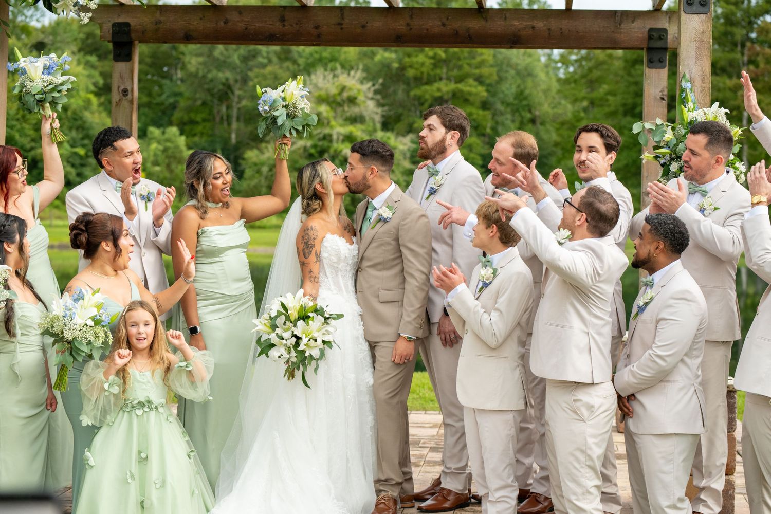 Wedding party celebrating outdoors under wooden arch, bridesmaids in mint green, groomsmen in tan suits, bride and groom kissing.