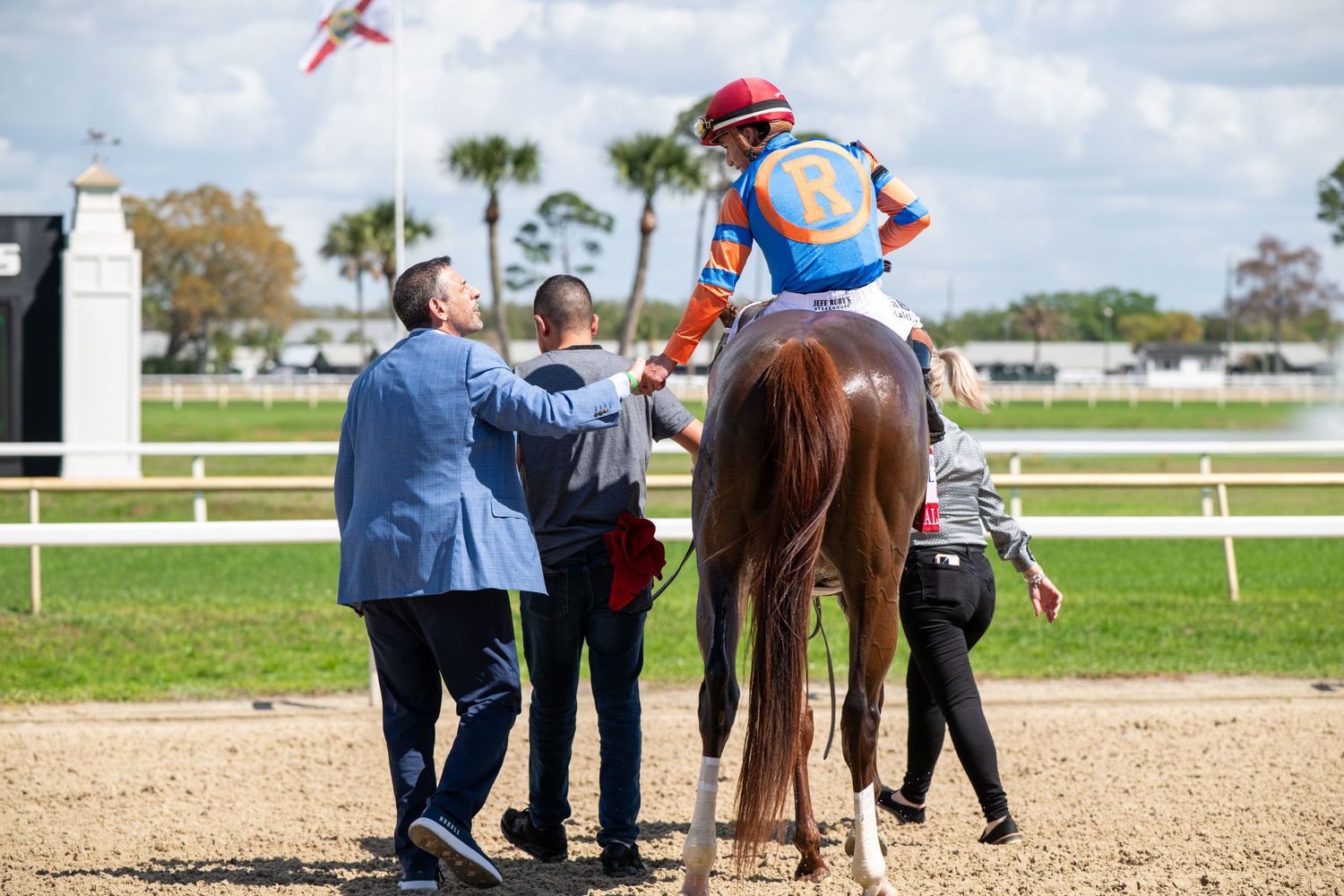 Jockey on a chestnut horse is assisted by handlers near palm trees at a Florida horse racing track.