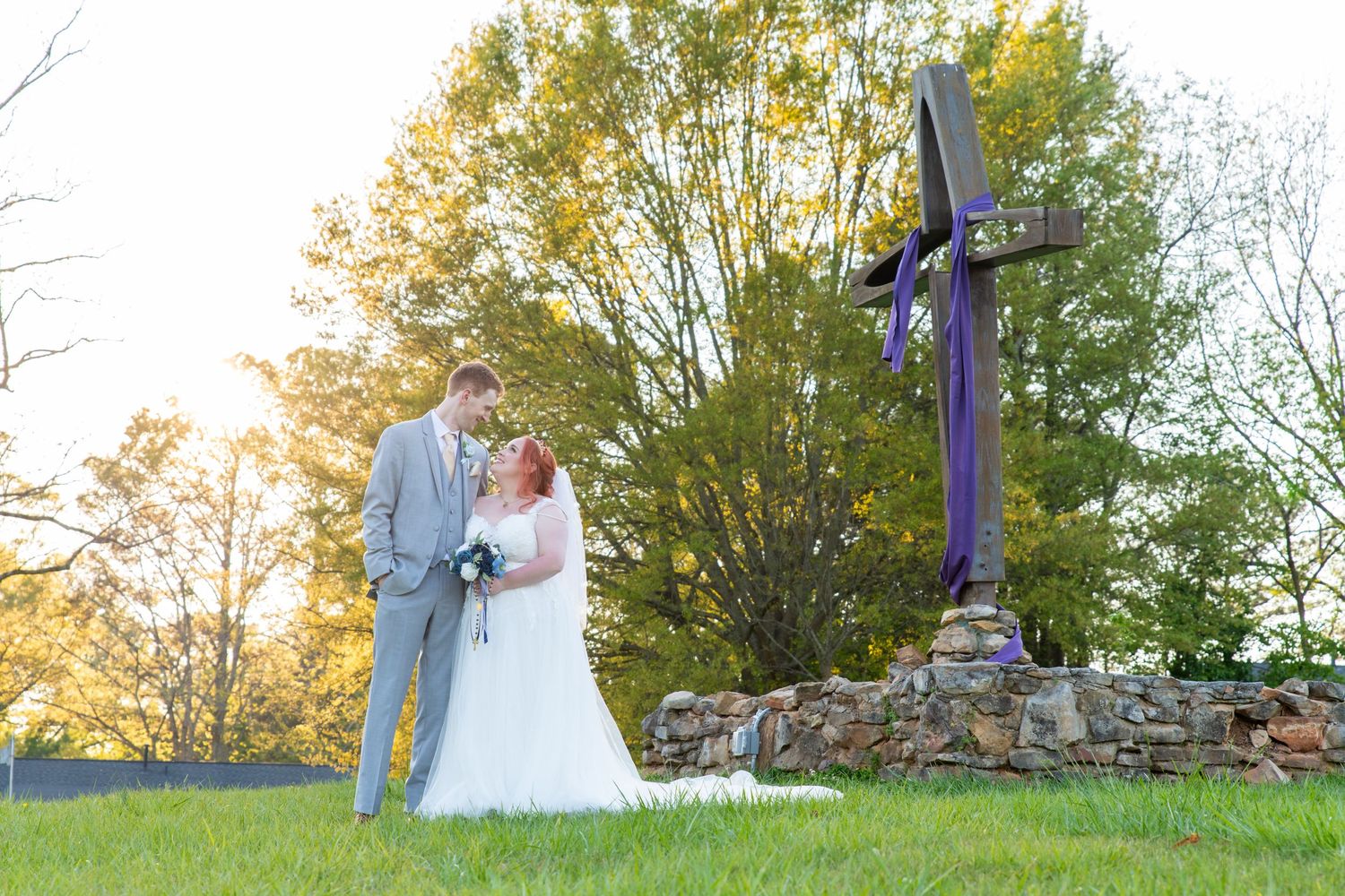 Bride and groom share a romantic moment beside a rustic stone cross at golden hour outdoor wedding.