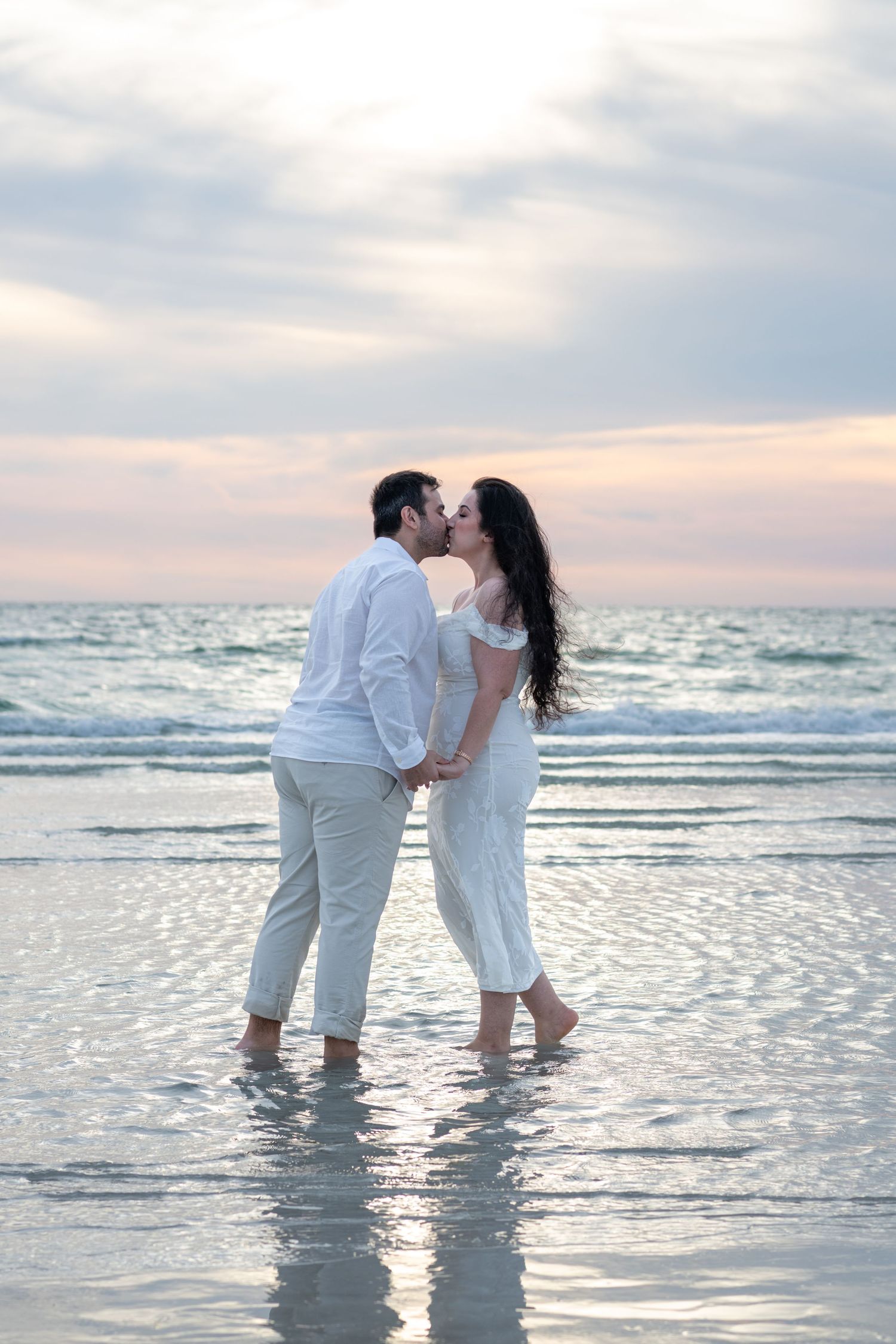 Couple kissing on beach at sunset, both wearing white, standing barefoot in shallow ocean water.