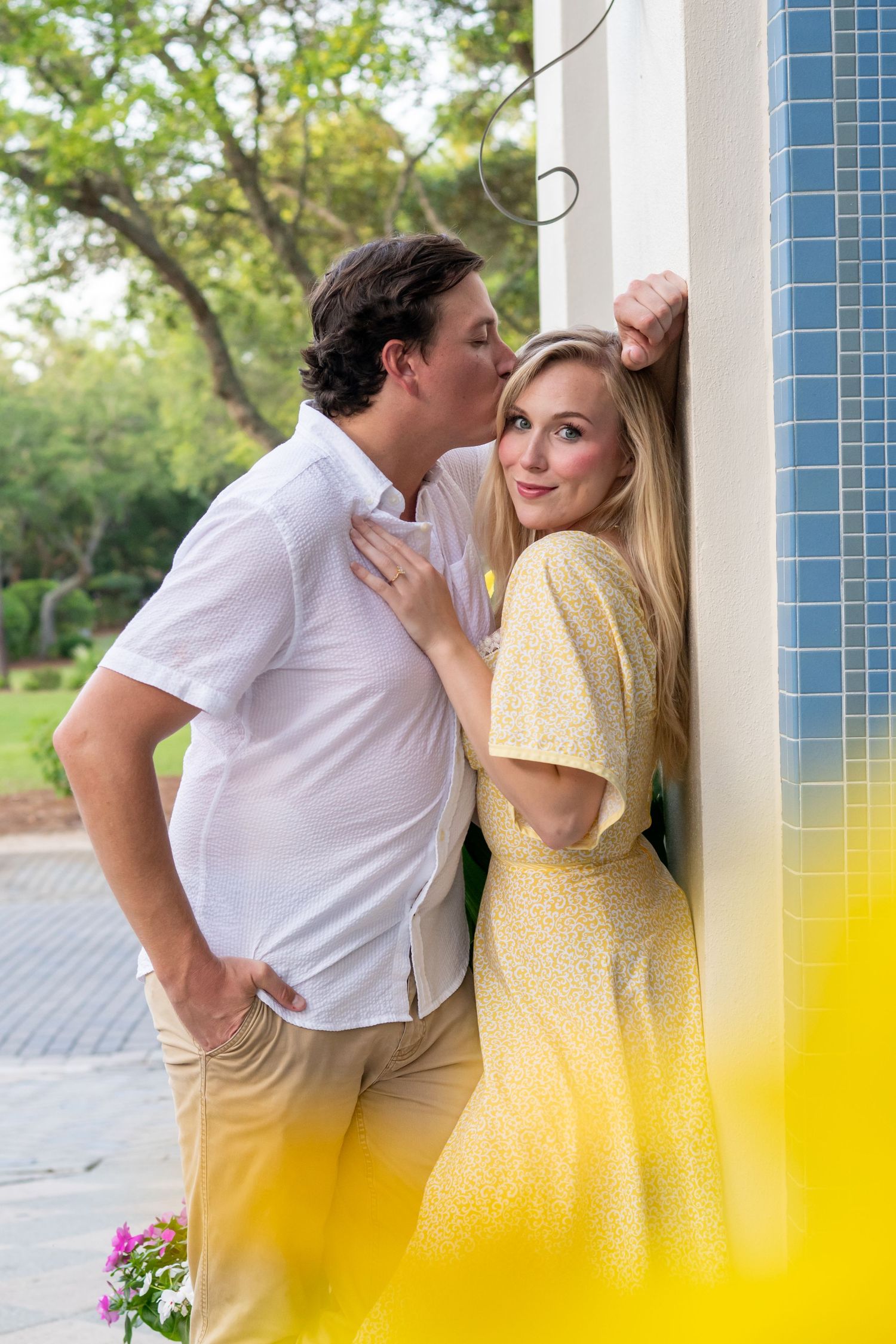 A man in white shirt kisses a smiling blonde woman in yellow dress leaning against a tiled wall outdoors.