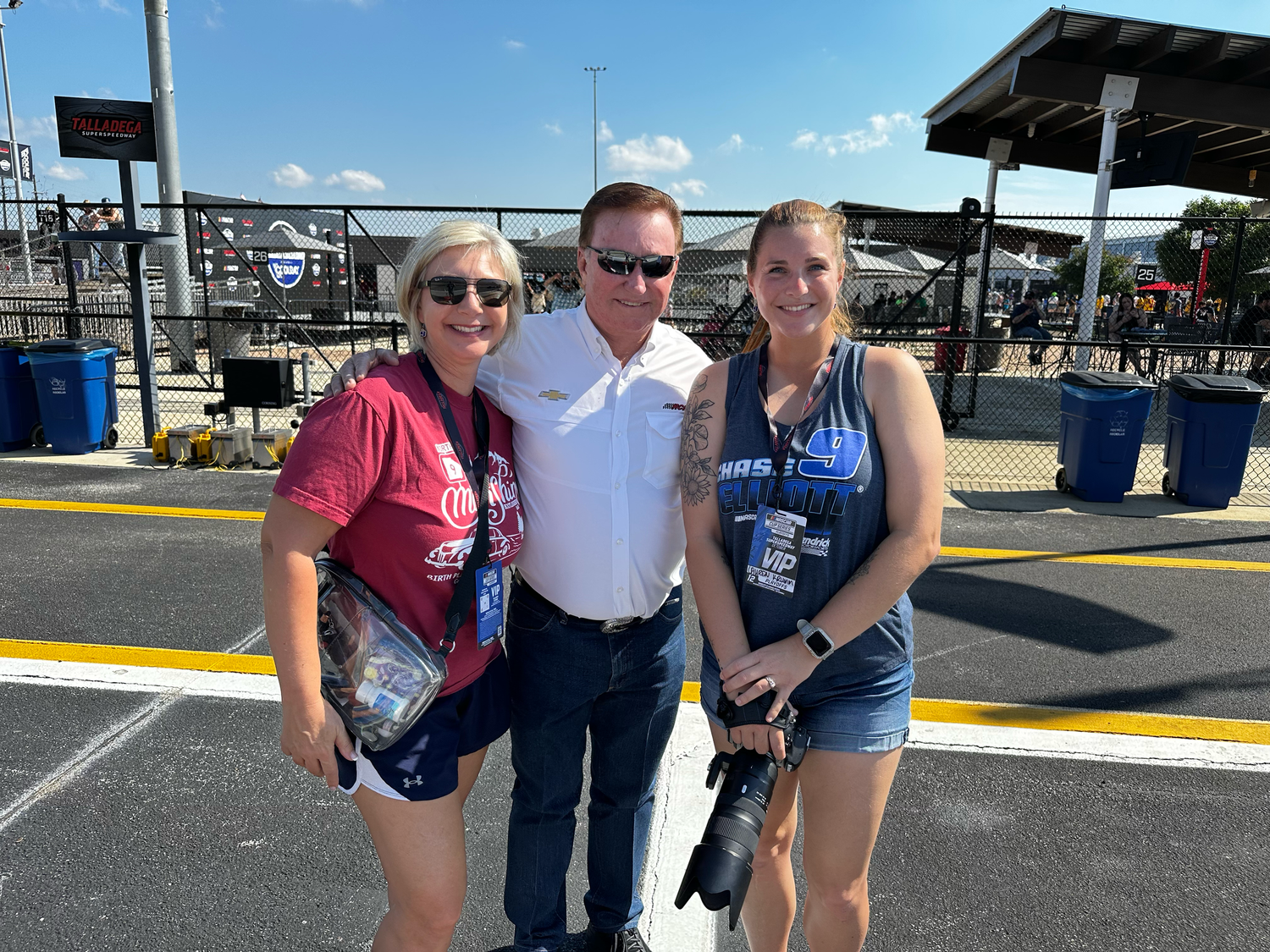 Three people posing together at a NASCAR racing event on a sunny day near the pit area.