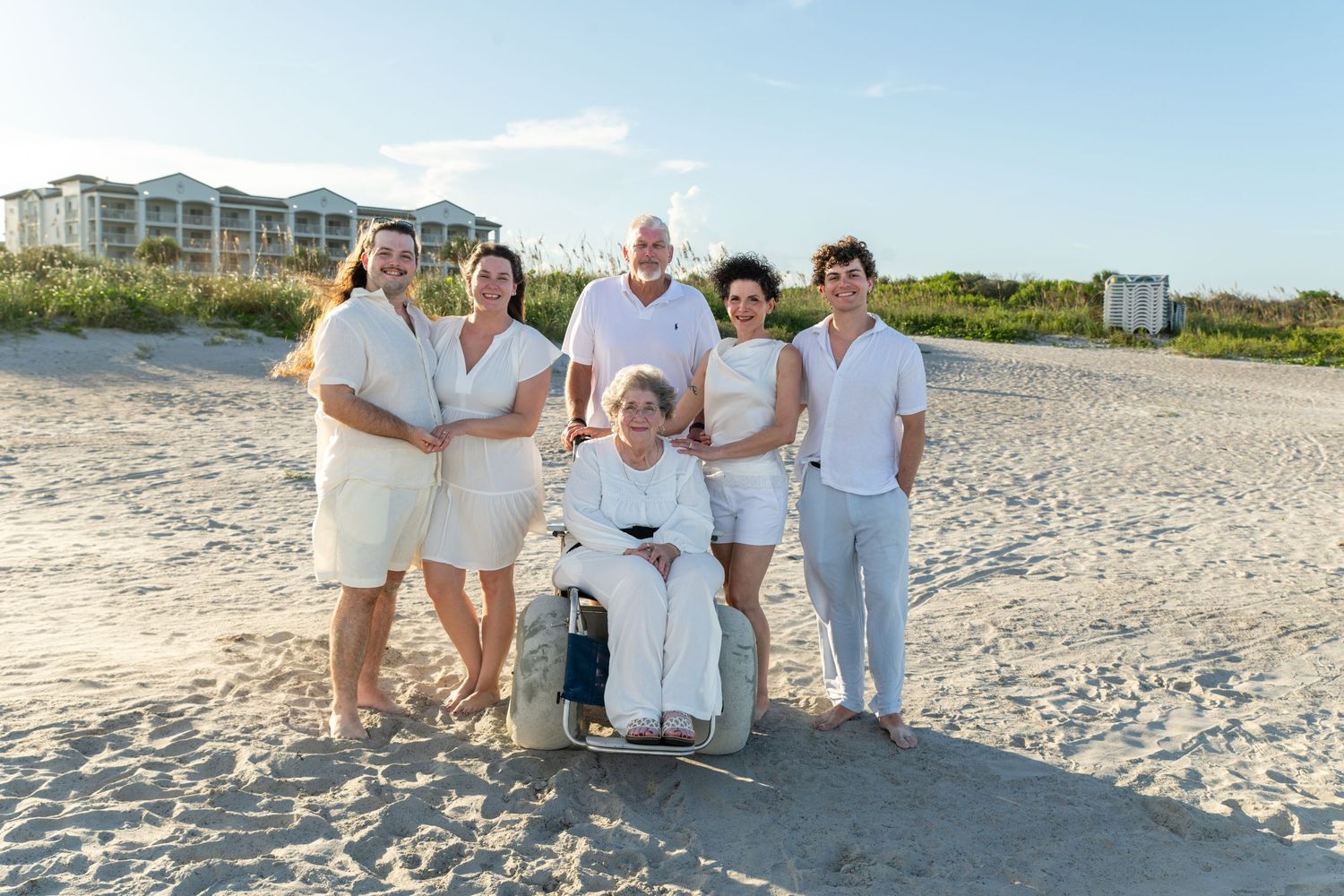 Family of six poses together on a sandy beach, one member in a wheelchair, all wearing white outfits at sunset.