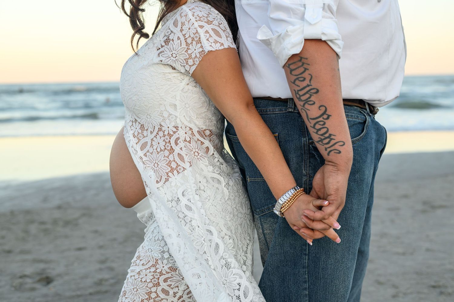 Pregnant woman in a white lace dress embraces her baby bump while a tattooed man holds her hand on a beach at sunset.