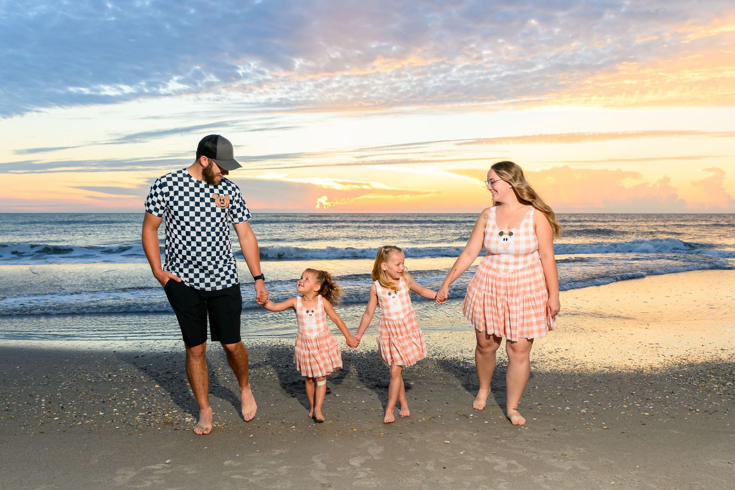 Family of four in matching coral outfits holding hands and walking along the beach at sunset with colorful sky and ocean waves.