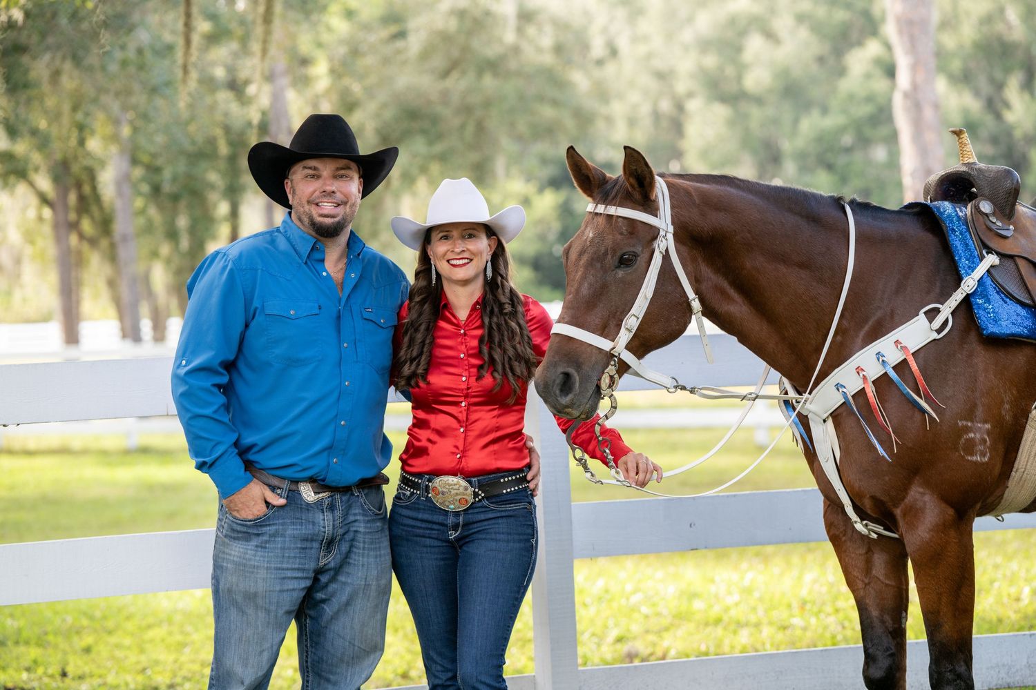 Couple in western attire posing with a chestnut horse by a white fence outdoors.