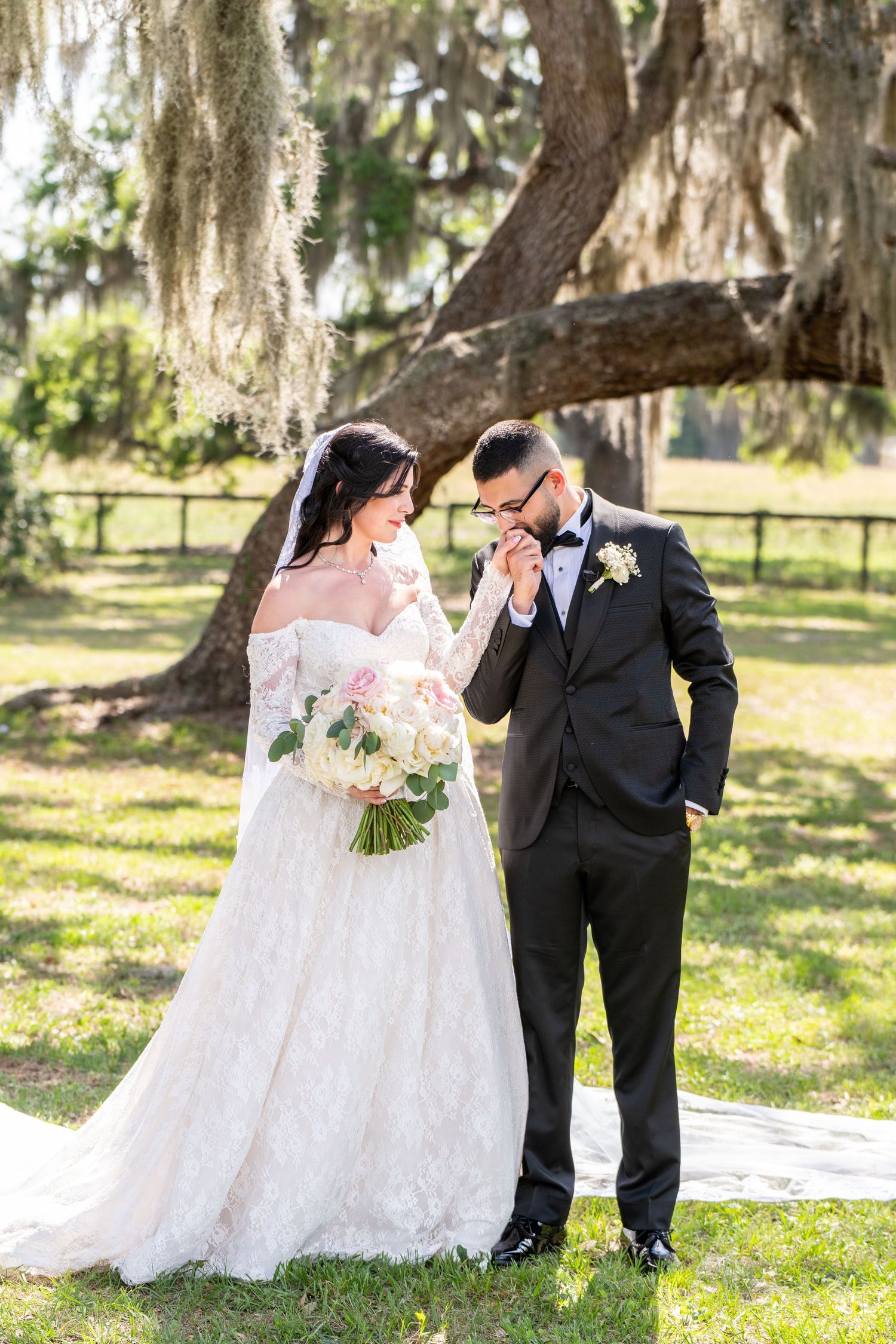 Groom in black tuxedo kissing bride's hand as she smiles in an off-shoulder lace wedding gown outdoors.