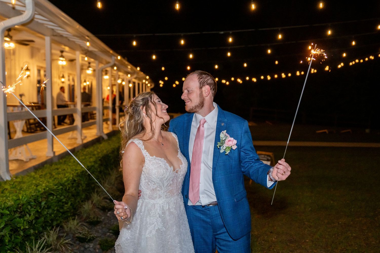 Bride and groom hold sparklers and share a romantic glance outside lit wedding venue at night.