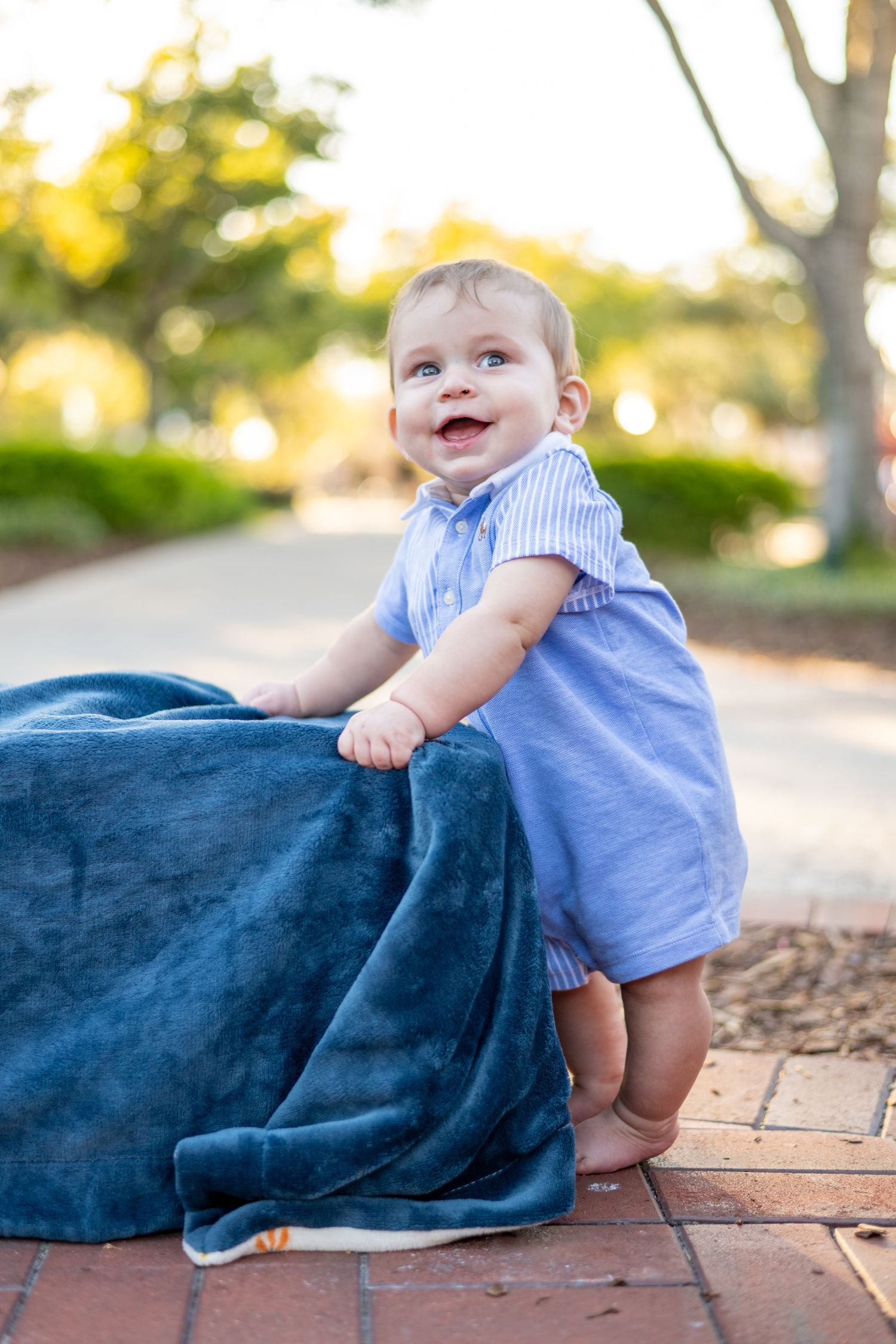 Happy baby in a blue romper standing and holding a navy blanket on a brick path.