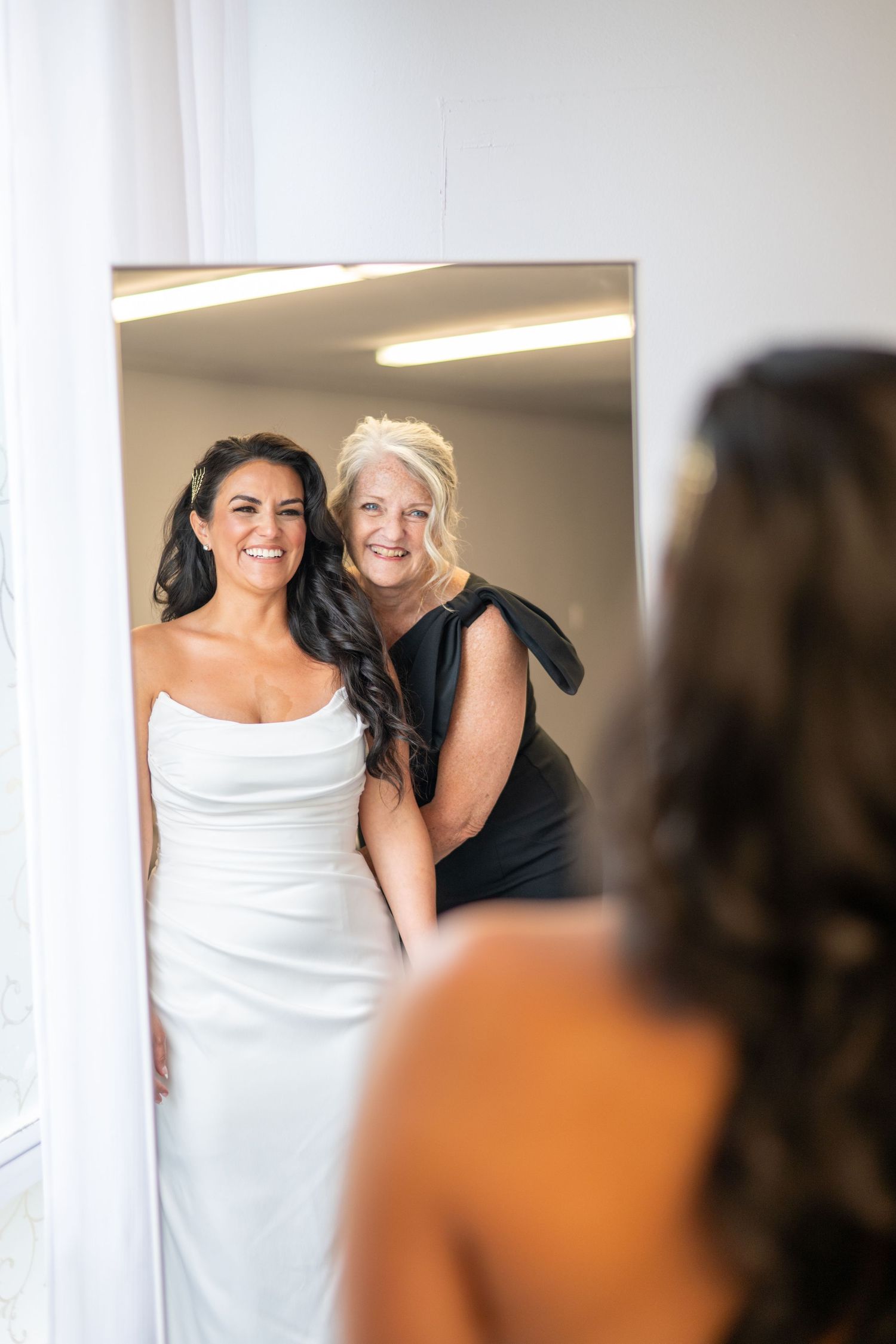 Bride in white strapless gown smiles with older woman in black dress, reflected in mirror during wedding preparations.