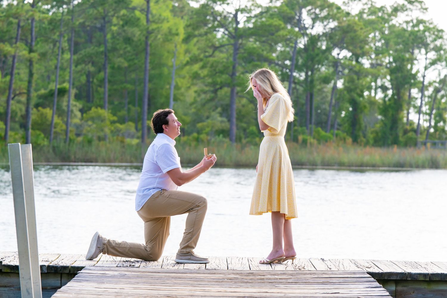Man kneeling on a dock proposing to a woman in a yellow dress by a serene lake surrounded by trees.