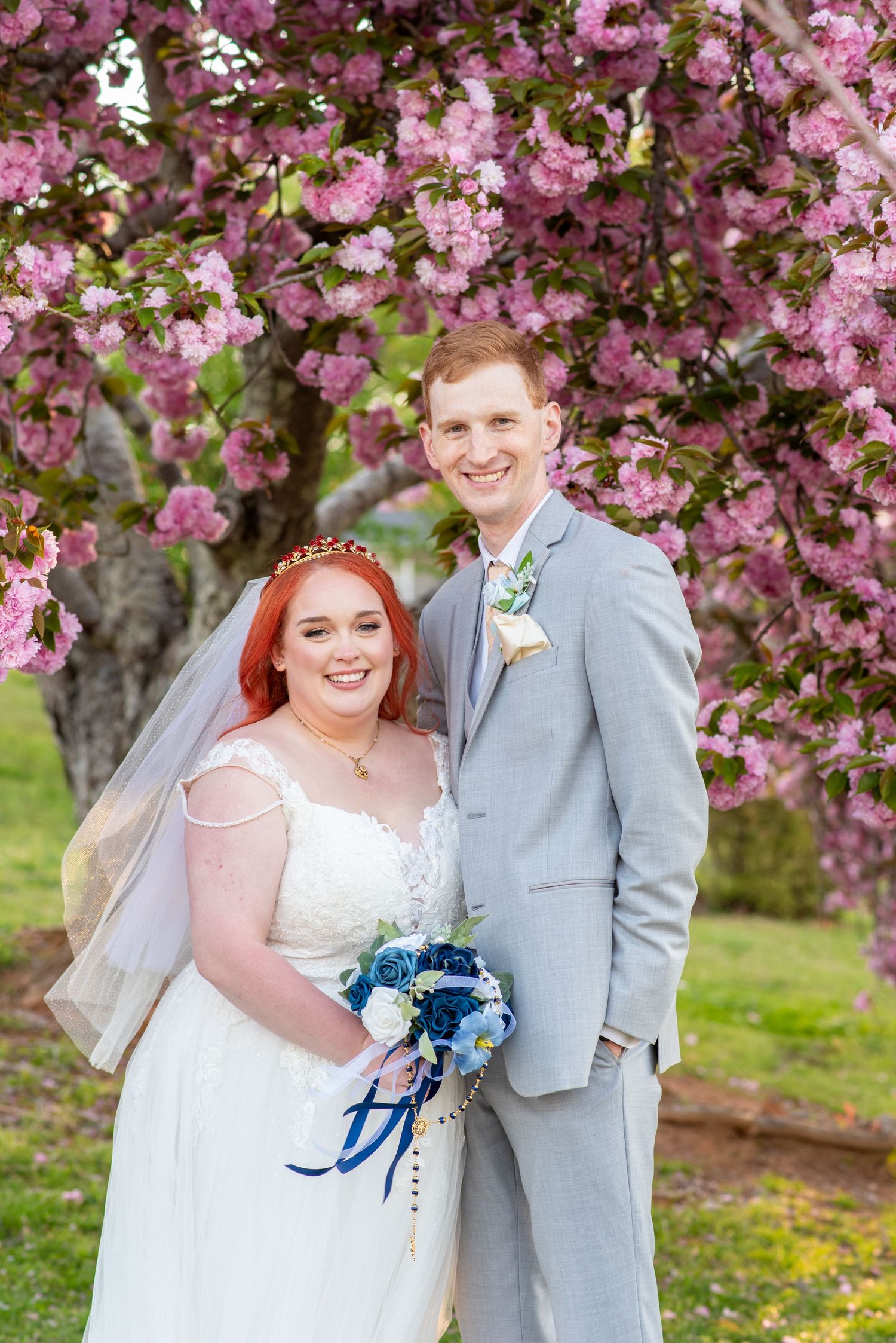 Bride and groom smiling under pink cherry blossom tree, bride holding blue bouquet in white wedding dress.