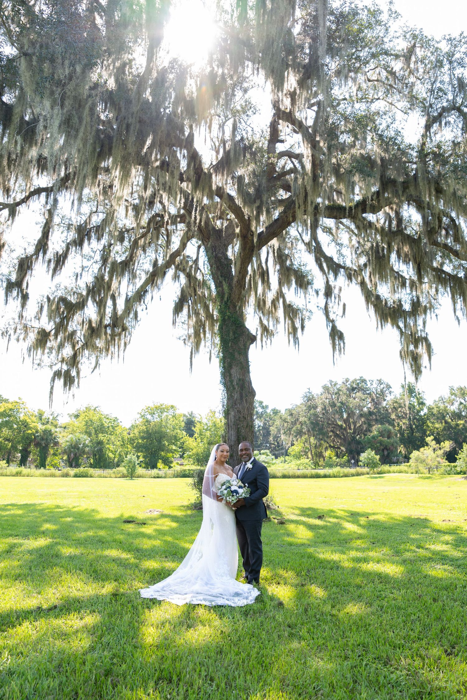 Bride and groom pose under a moss-draped oak tree on a lush green lawn during their outdoor wedding ceremony.