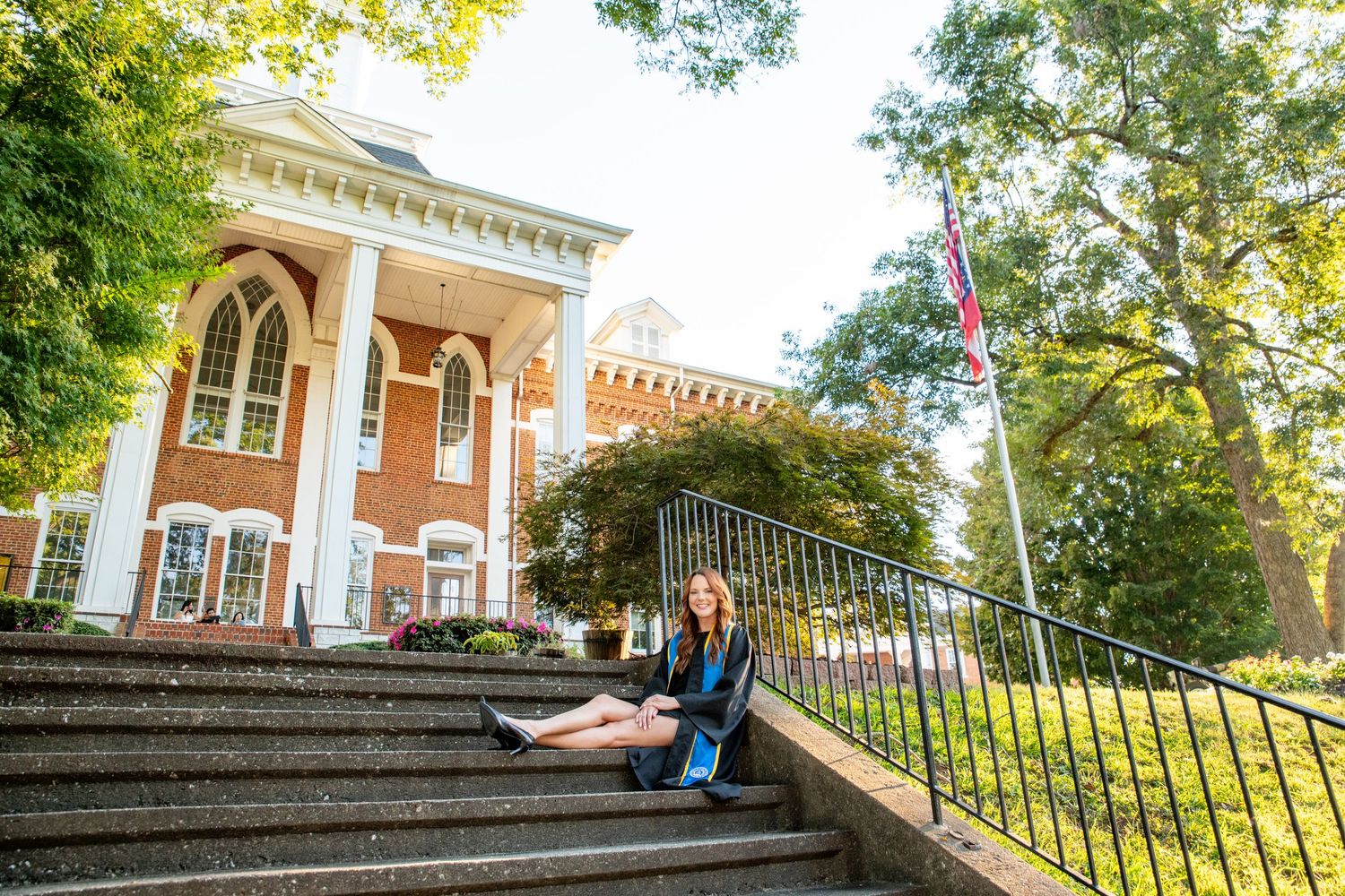 A young woman in graduation attire sits on brick steps outside a red brick university building with a flagpole nearby.