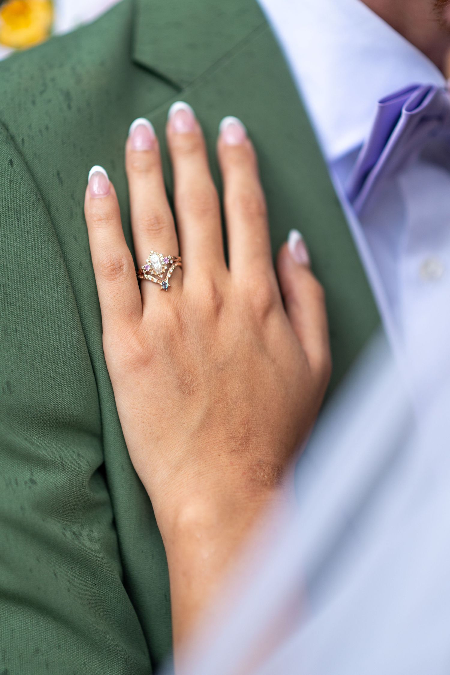 Close-up of bride's hand with elegant gold crown wedding ring resting on groom's green suit jacket.