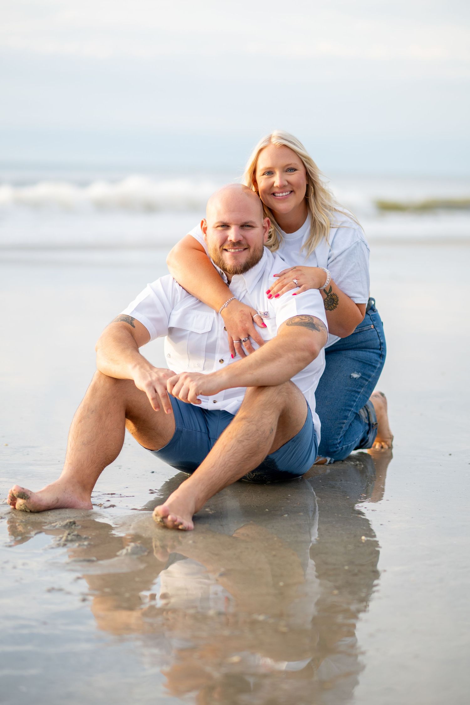 Couple sitting on wet beach sand, woman hugging man from behind, both wearing white shirts and jeans.