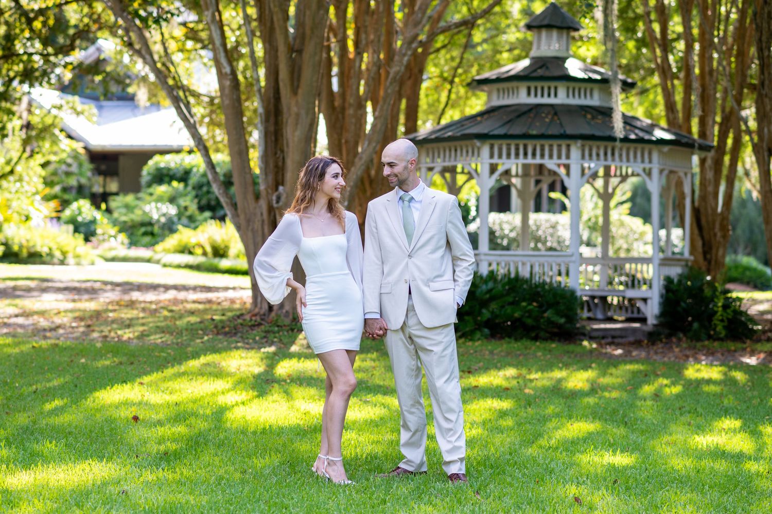 Couple holding hands smiling at each other in front of a white gazebo on a lush green lawn.