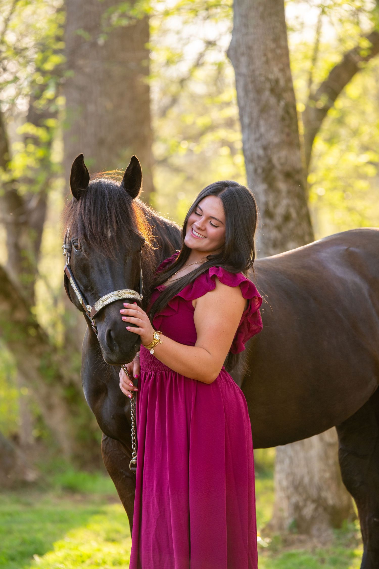 Woman in magenta dress smiling while embracing a dark brown horse in a sunlit forest setting.
