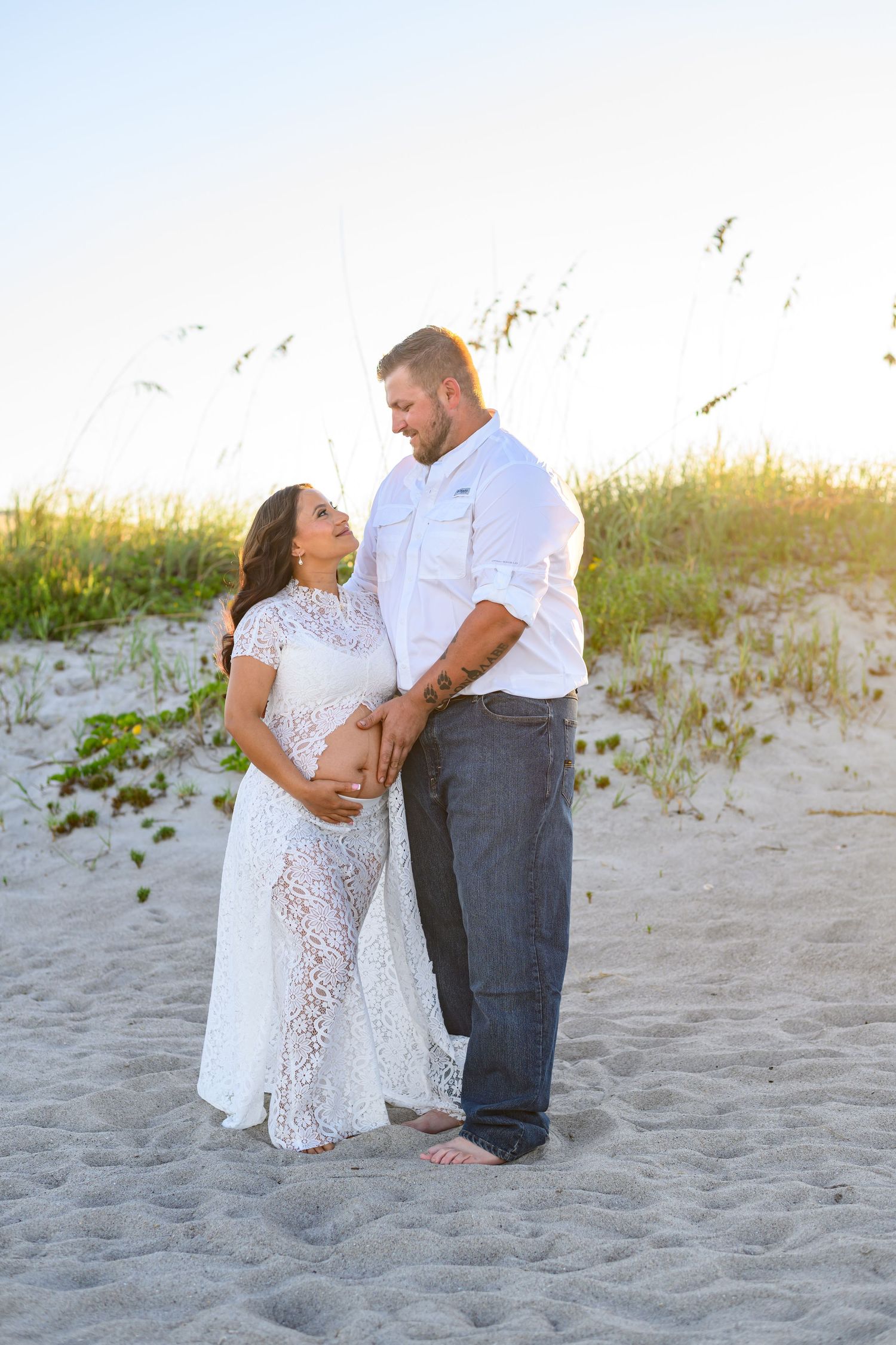 Expecting couple embracing on a sandy beach at sunset, the woman in a white lace maternity gown and the man in a white shirt and jeans.