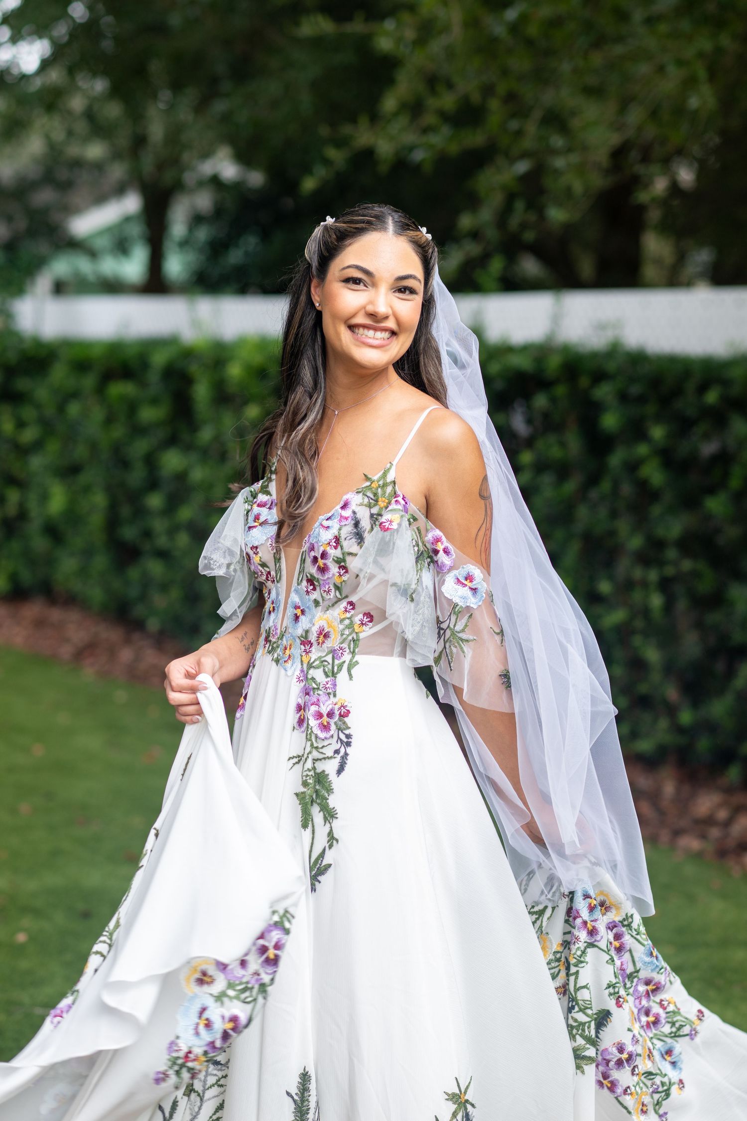 Smiling bride in floral embroidered white wedding gown with veil, posing outdoors in lush garden setting.