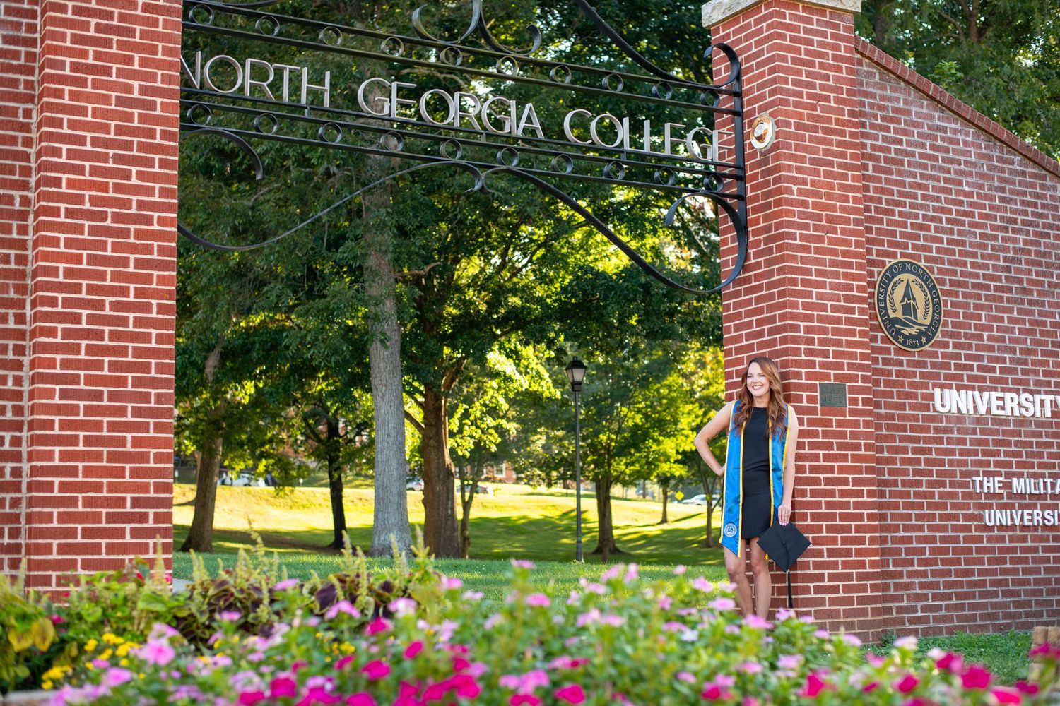 Graduate in cap and gown poses under North Georgia College University brick entrance arch surrounded by colorful flowers.