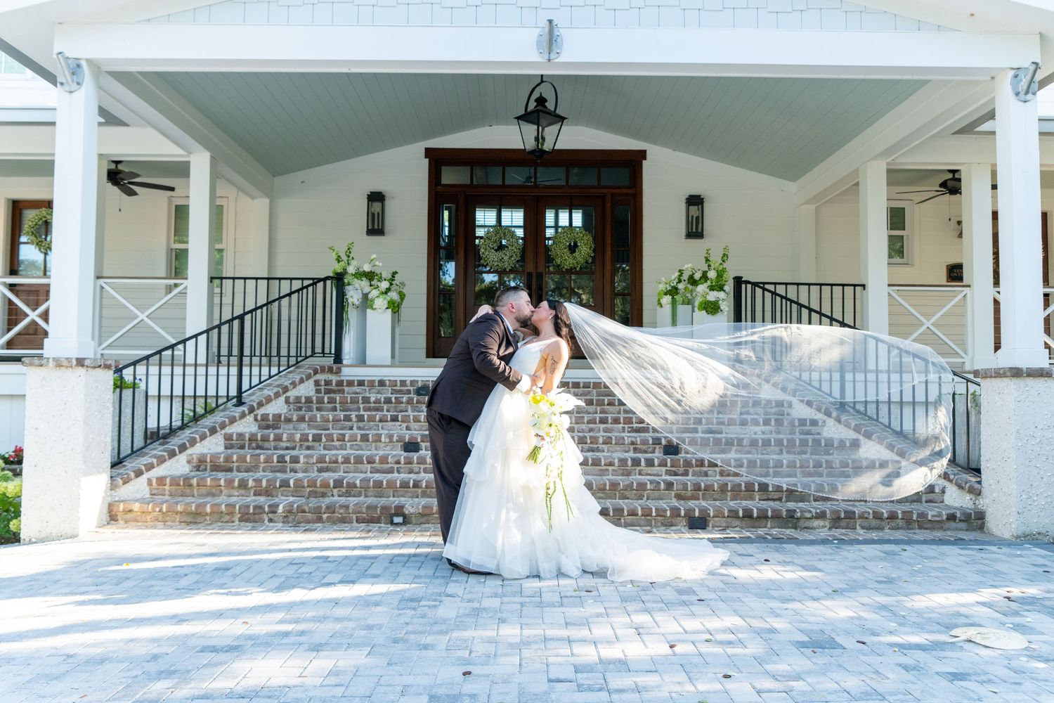 Bride and groom share a romantic kiss on grand staircase steps outside elegant wedding venue with veil flowing in wind.