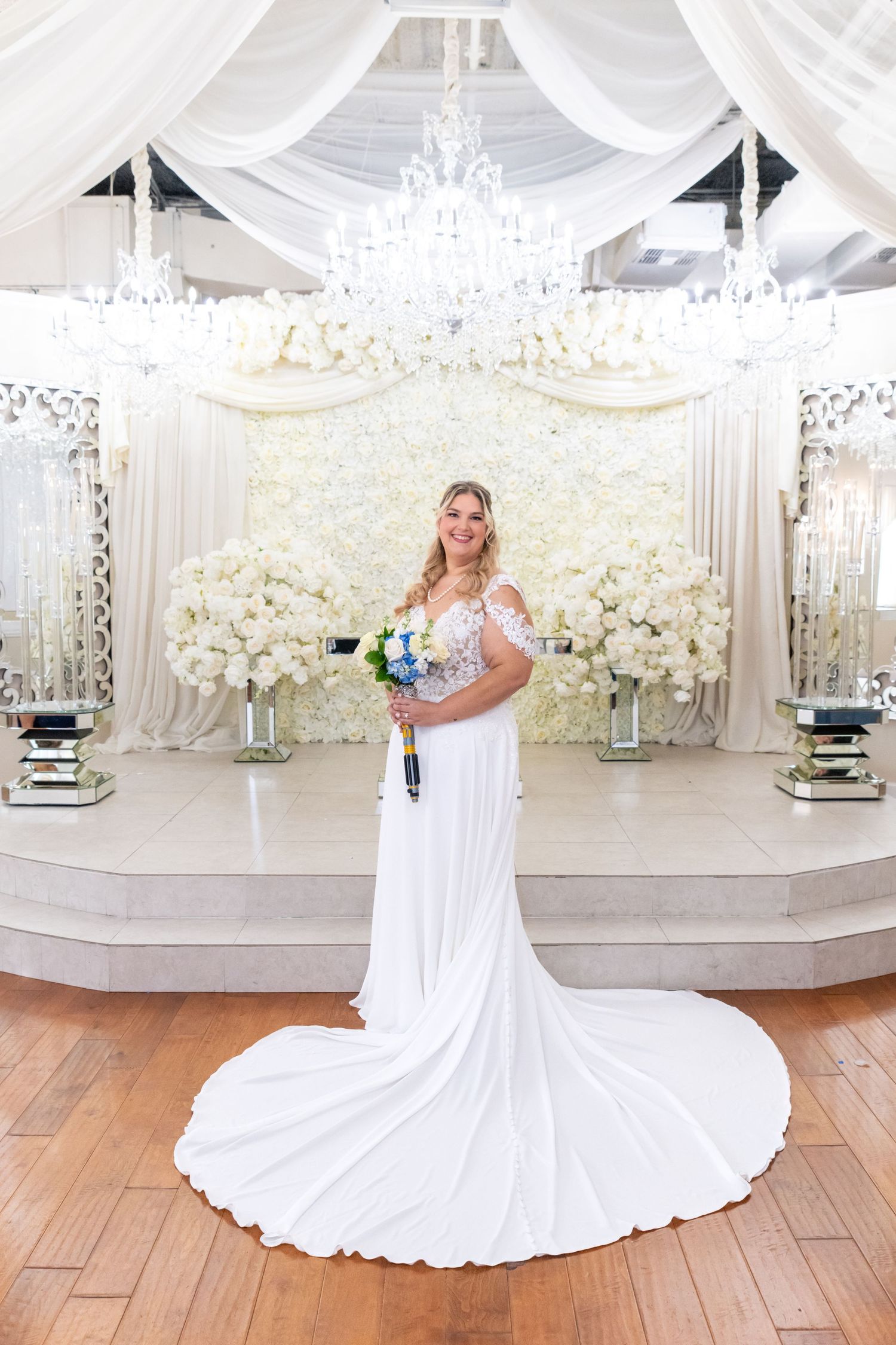 Bride in white gown with long train holds bouquet before floral backdrop with chandeliers in elegant wedding venue.