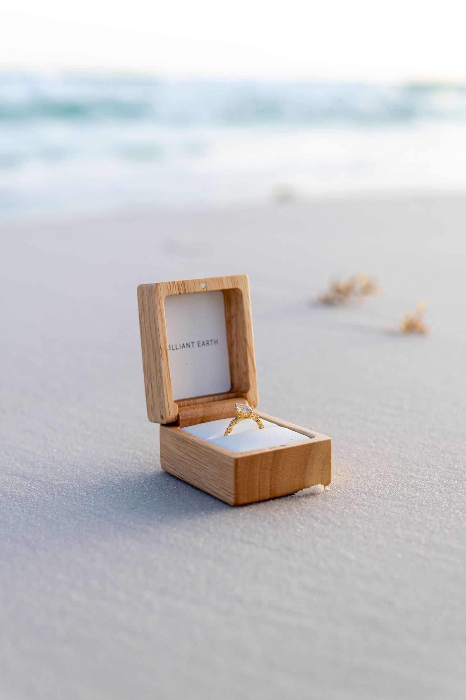 Gold diamond engagement ring in wooden Brilliant Earth box on white sand beach with ocean waves in background.