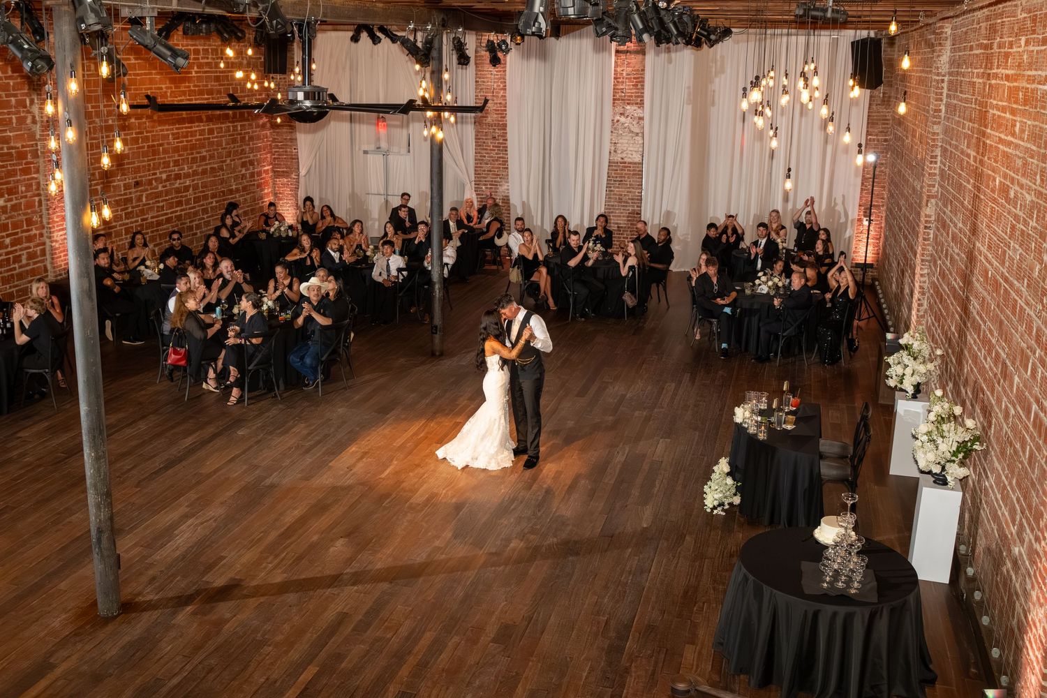 Bride and groom share first dance on hardwood floor in rustic brick venue with string lights and seated wedding guests.