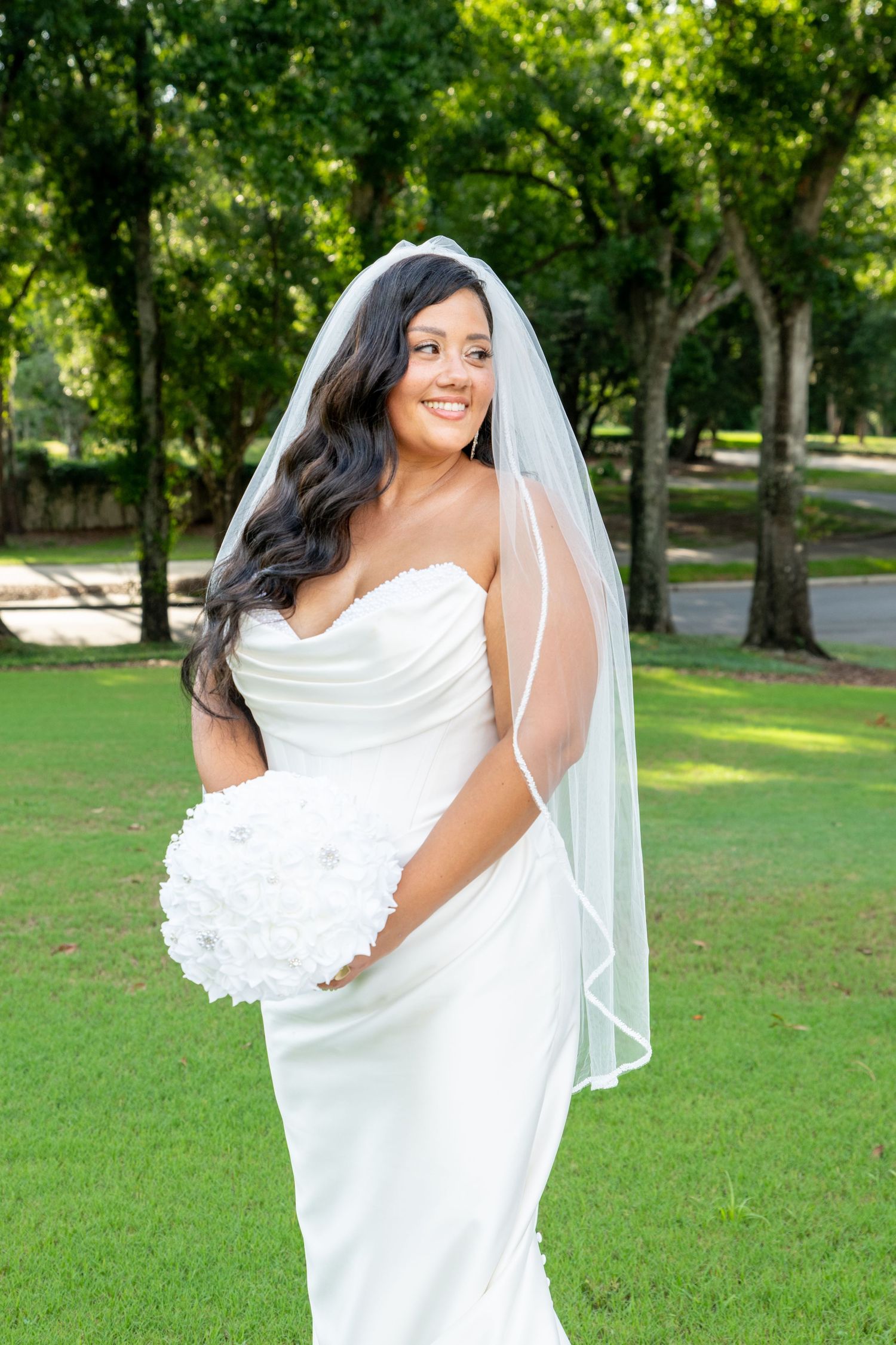 Smiling bride in strapless white gown and veil holds white bouquet outdoors on green lawn with trees.