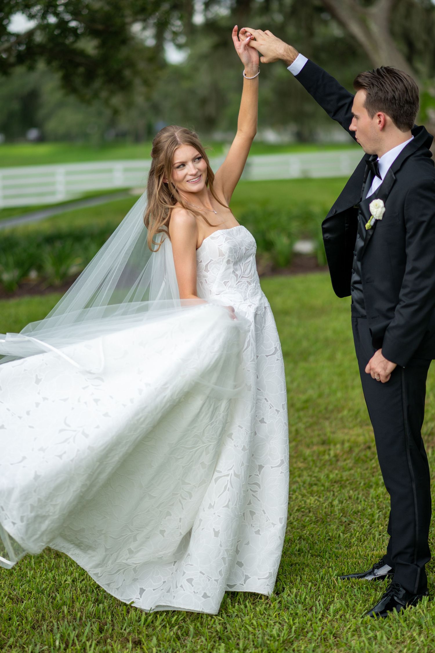 Groom twirling bride in white lace gown on green lawn during wedding photos.