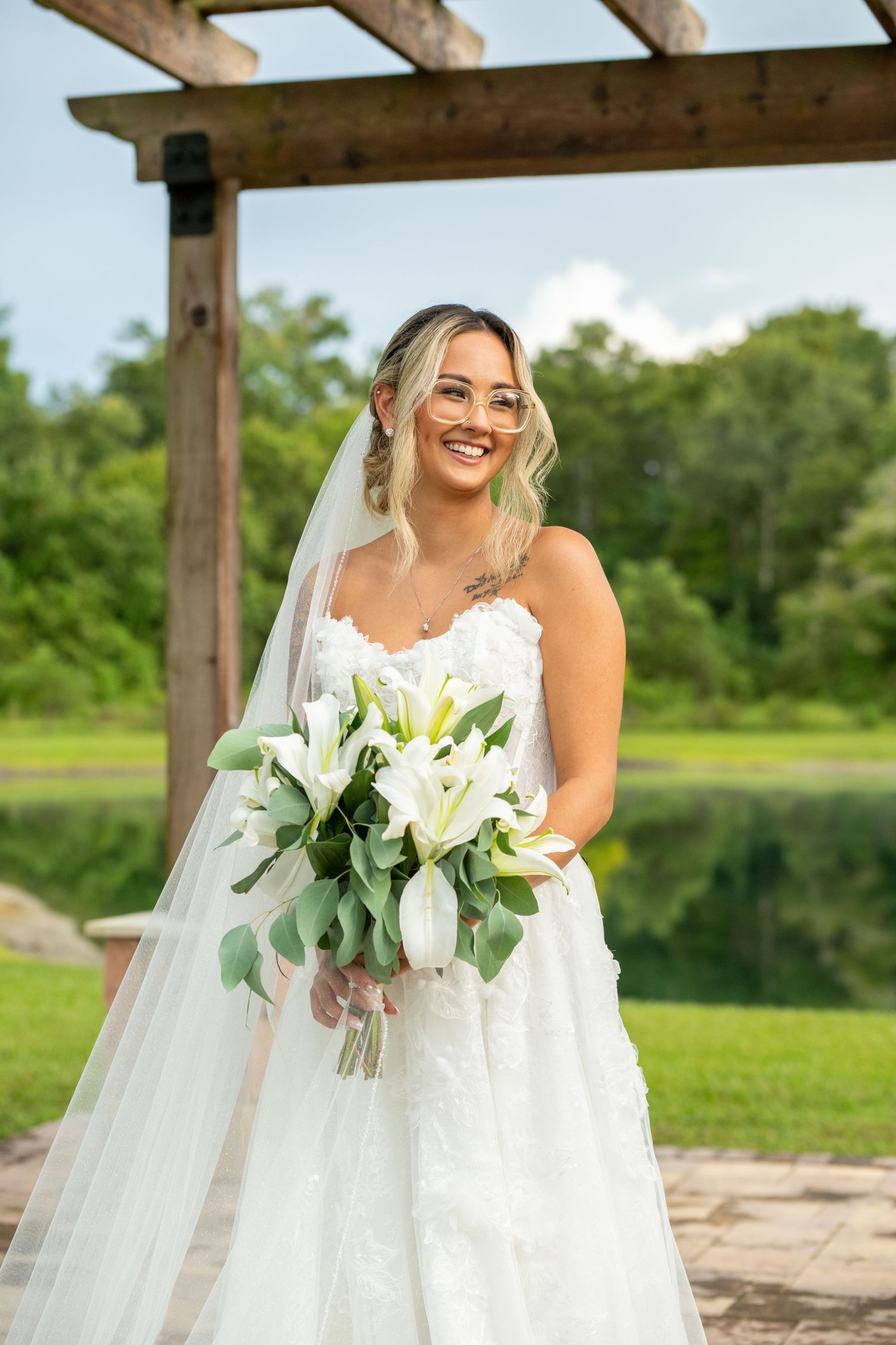 Smiling bride in white lace gown and veil holds white lily bouquet outdoors under wooden pergola.