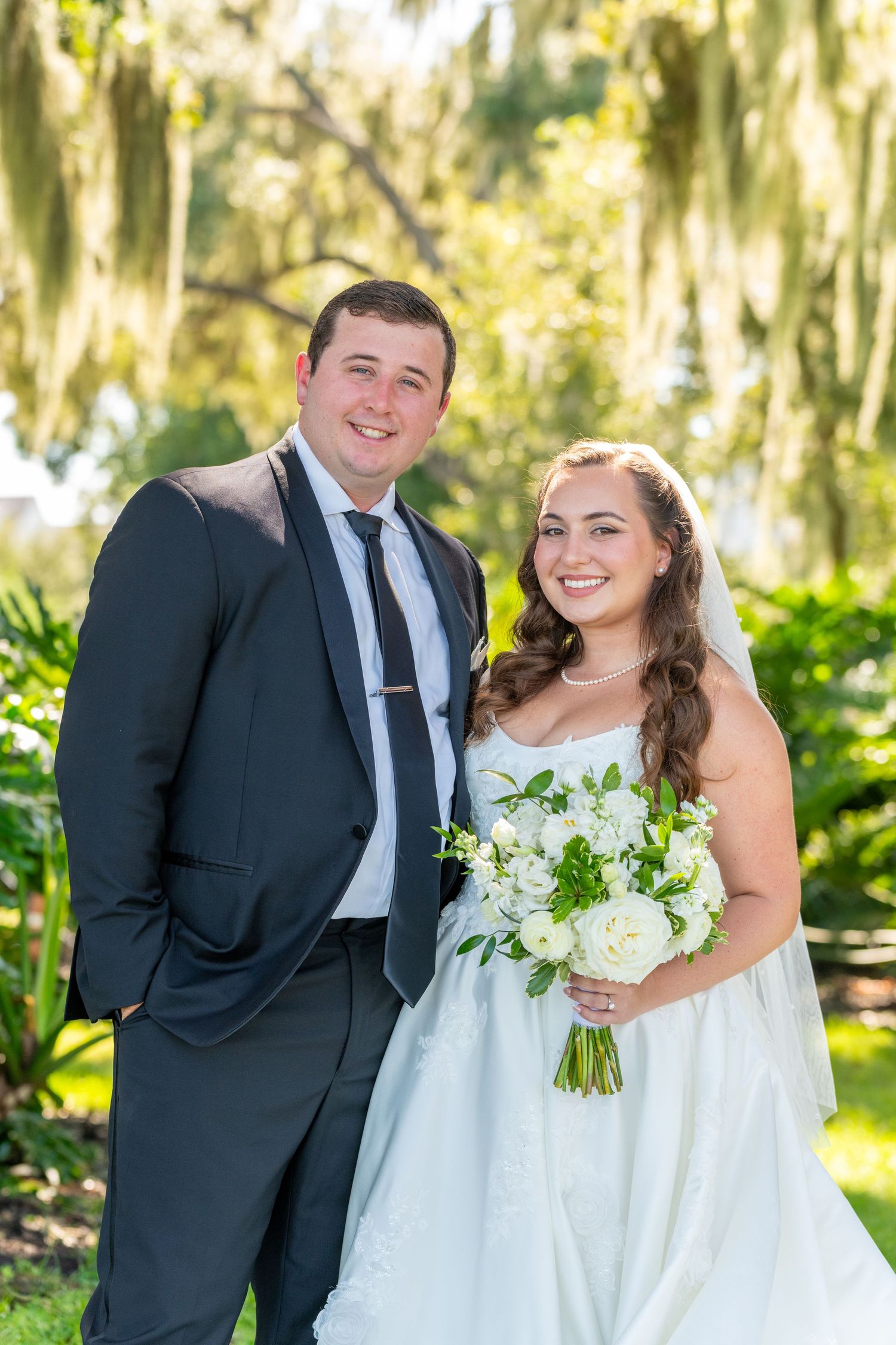 Bride and groom smiling outdoors under Spanish moss trees, bride holding white floral bouquet in elegant wedding attire.