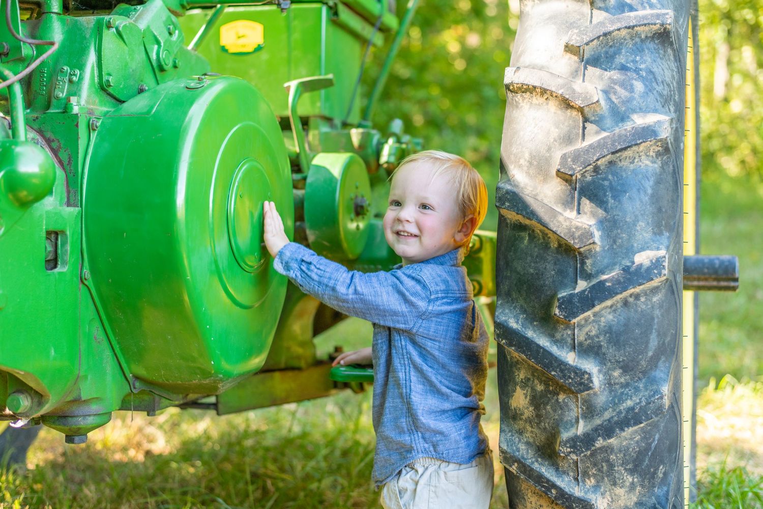 Happy toddler in denim shirt smiling while playing beside large green John Deere tractor tire.