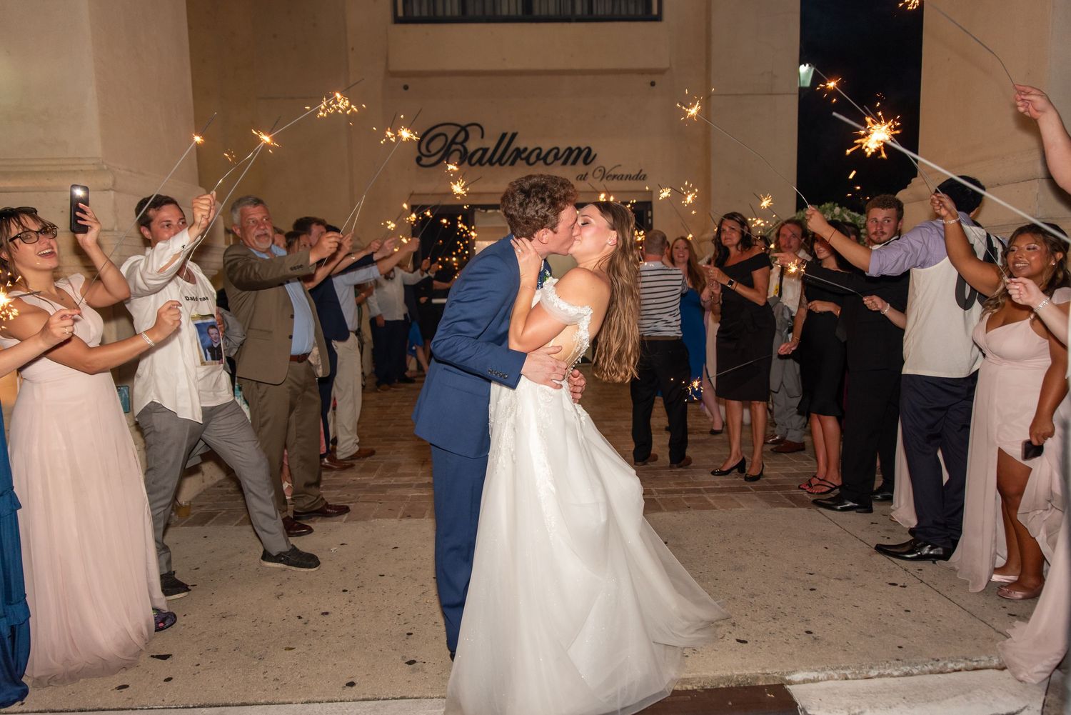 Bride and groom share passionate kiss during sparkler send-off outside ballroom entrance at night.