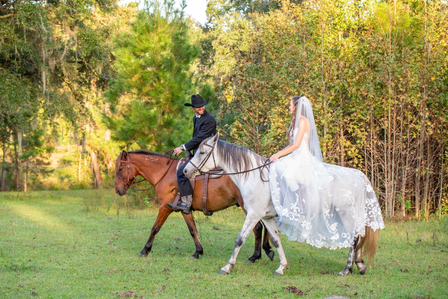Bride and groom riding horses through a lush green field in wedding attire, with bride's veil flowing behind.