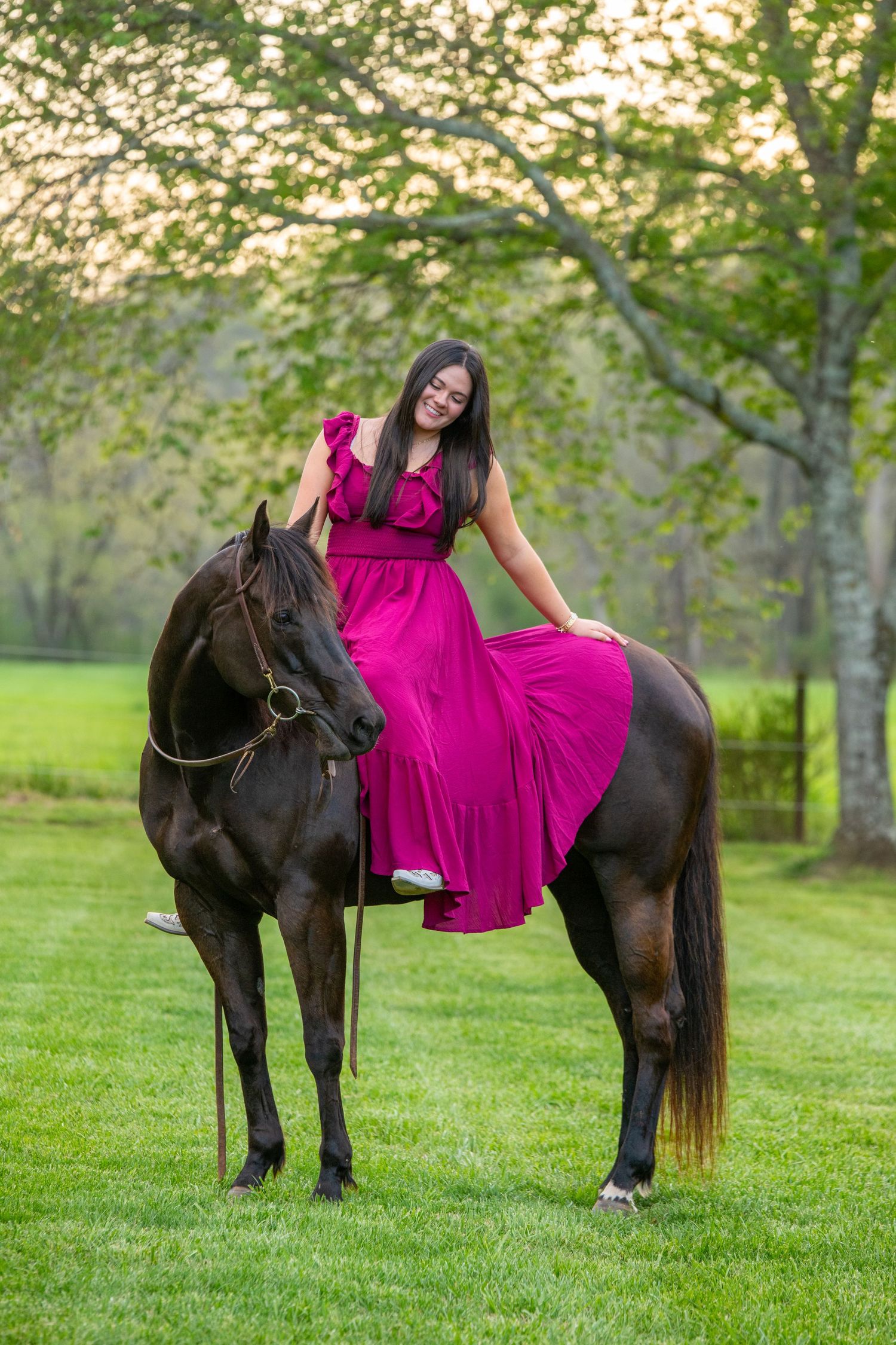 Woman in pink dress riding a dark horse on green grass with trees in background.