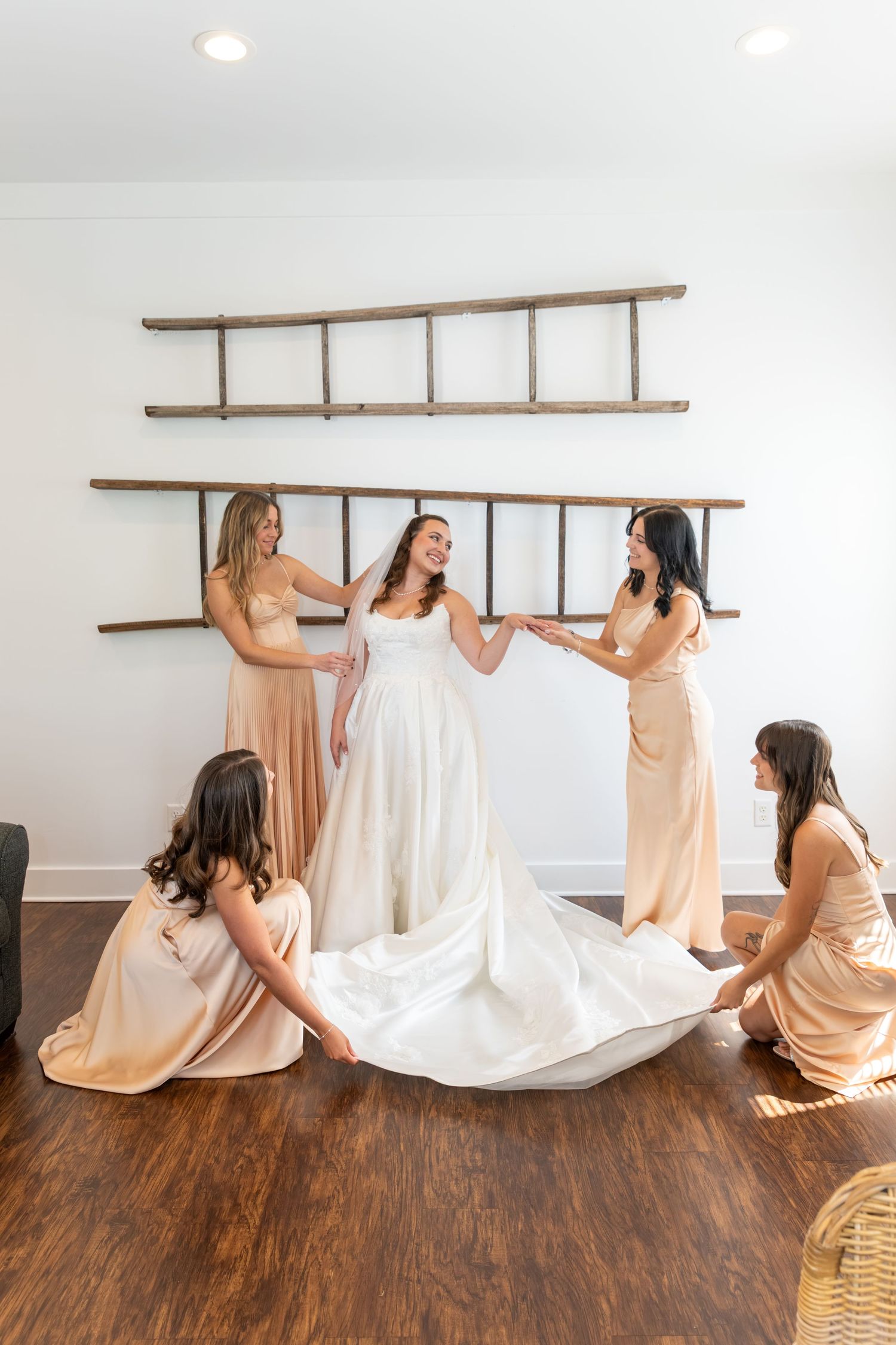 Bride in white gown surrounded by four bridesmaids in champagne dresses helping arrange her train indoors.