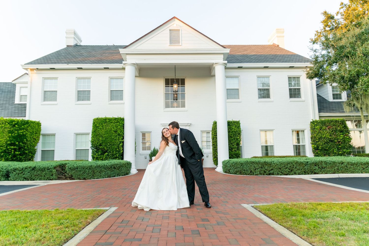 Bride and groom share a kiss in front of elegant white mansion with brick pathway and manicured hedges.