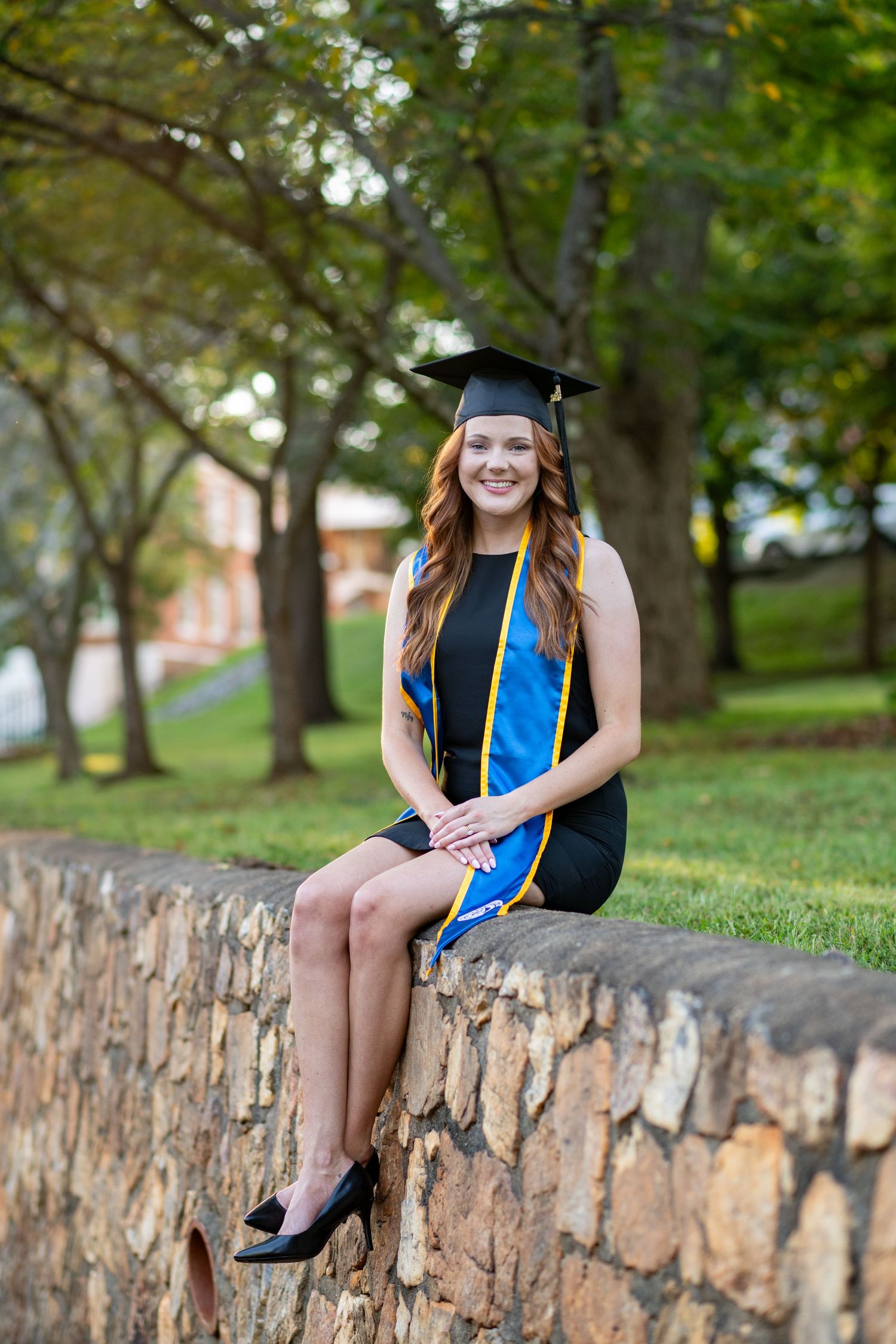 Smiling young woman in graduation cap and blue stole sits on stone wall outdoors in park setting.
