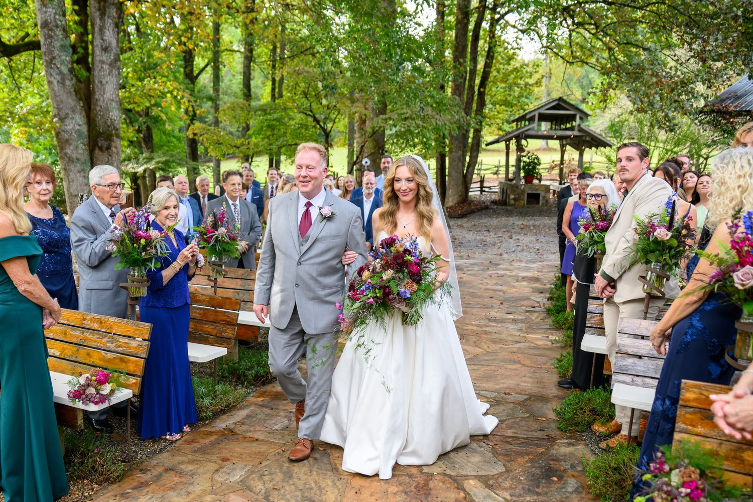 Bride and groom walk down outdoor aisle as guests toss flowers at rustic woodland wedding ceremony.