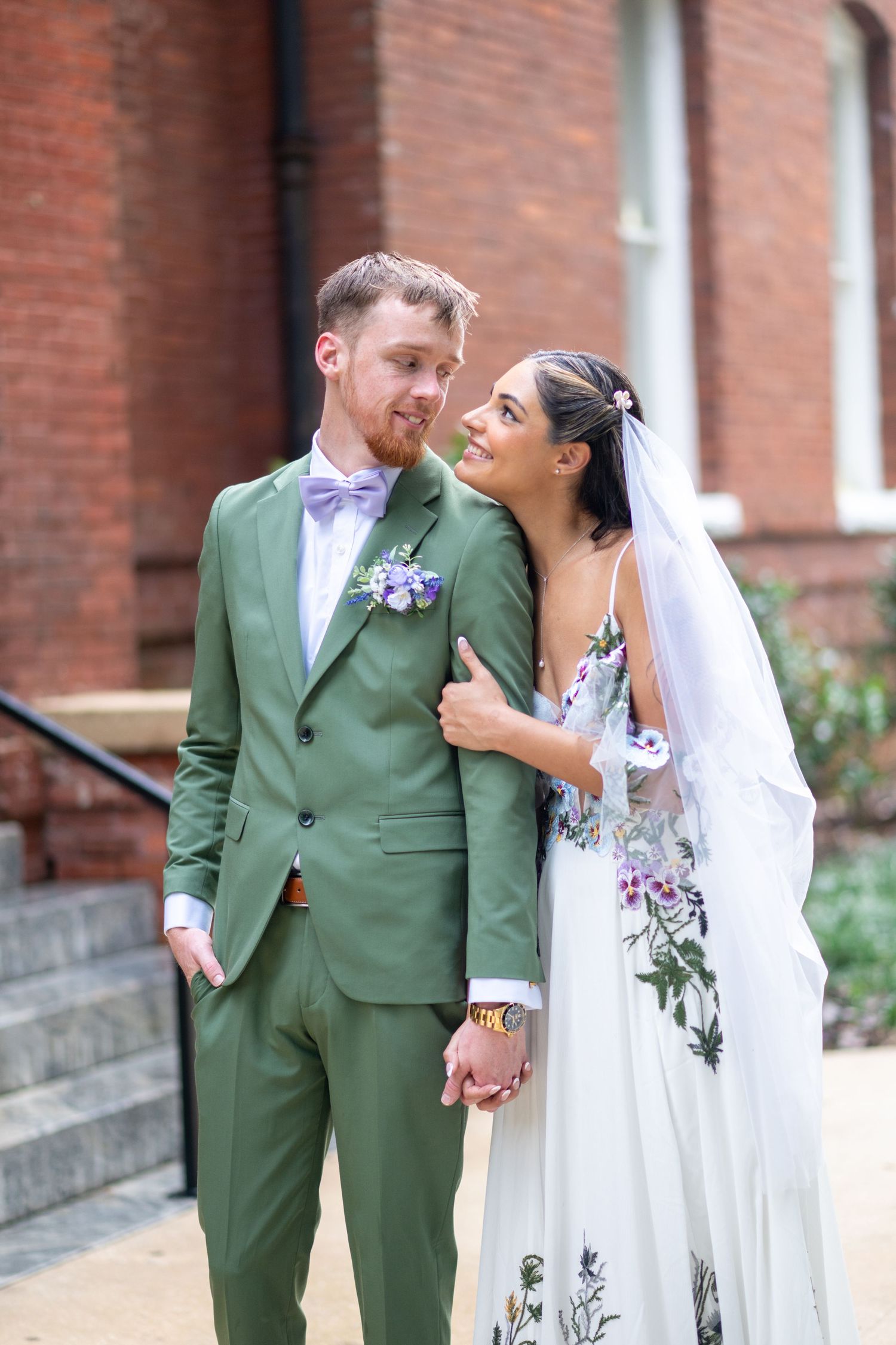 Bride in floral embroidered gown and groom in green suit with lavender bow tie share a tender moment outdoors.