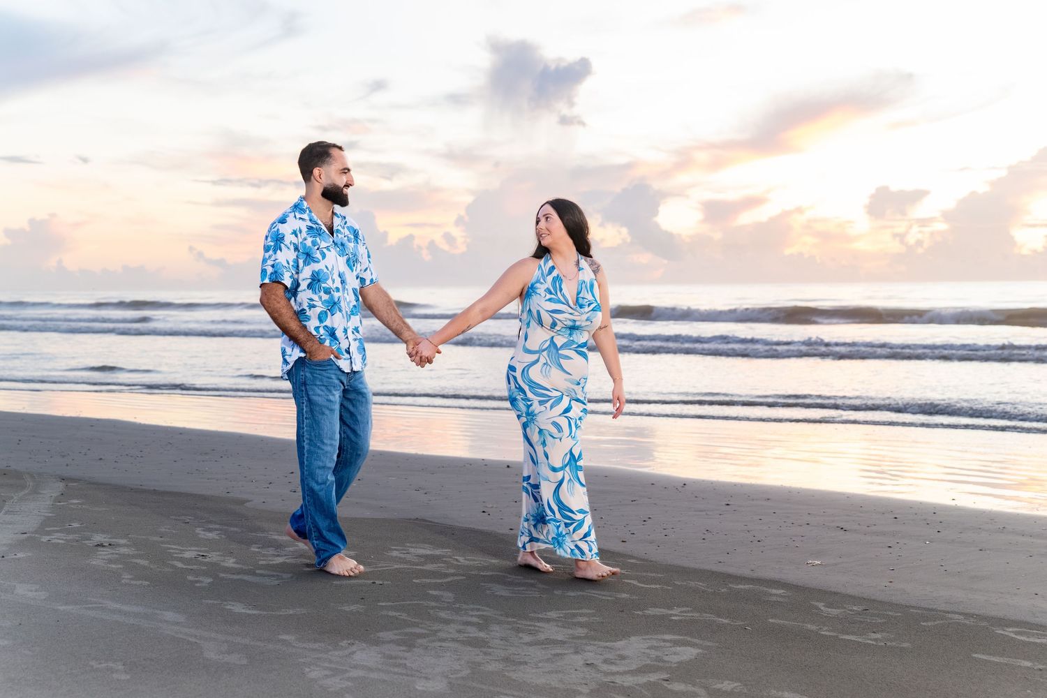 Couple holding hands on beach at sunset, wearing matching blue floral outfits, waves and pastel sky in background.