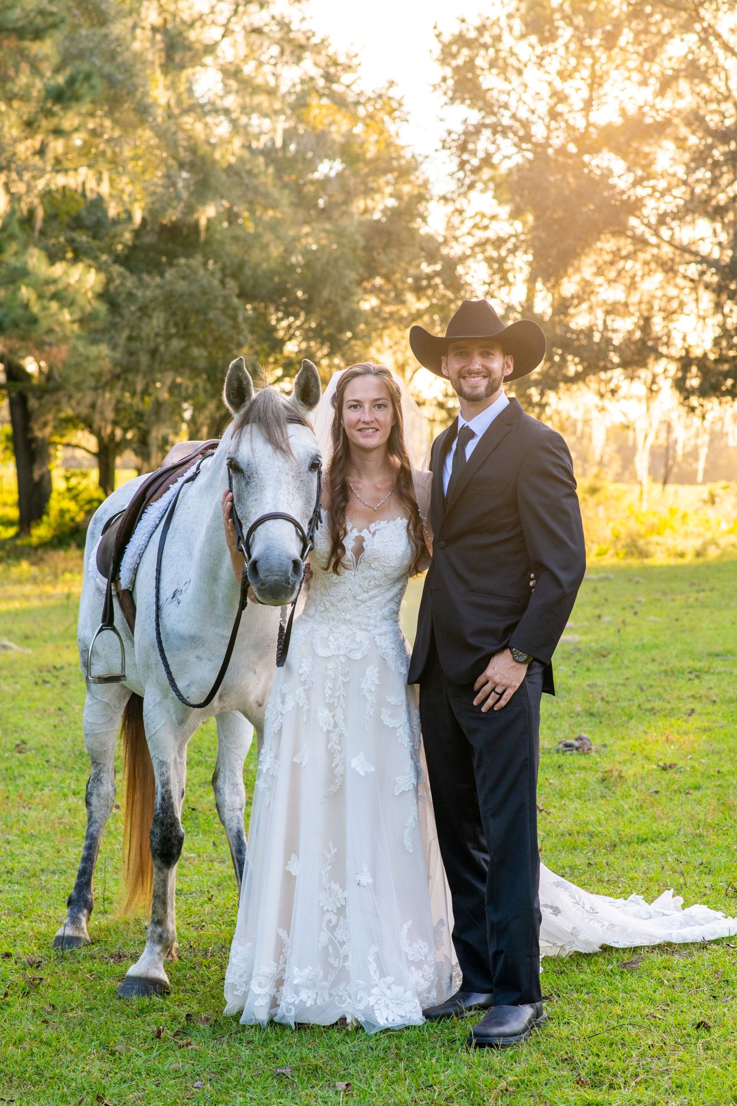 Bride and groom in cowboy hat posing with white horse at golden hour outdoor wedding ceremony.