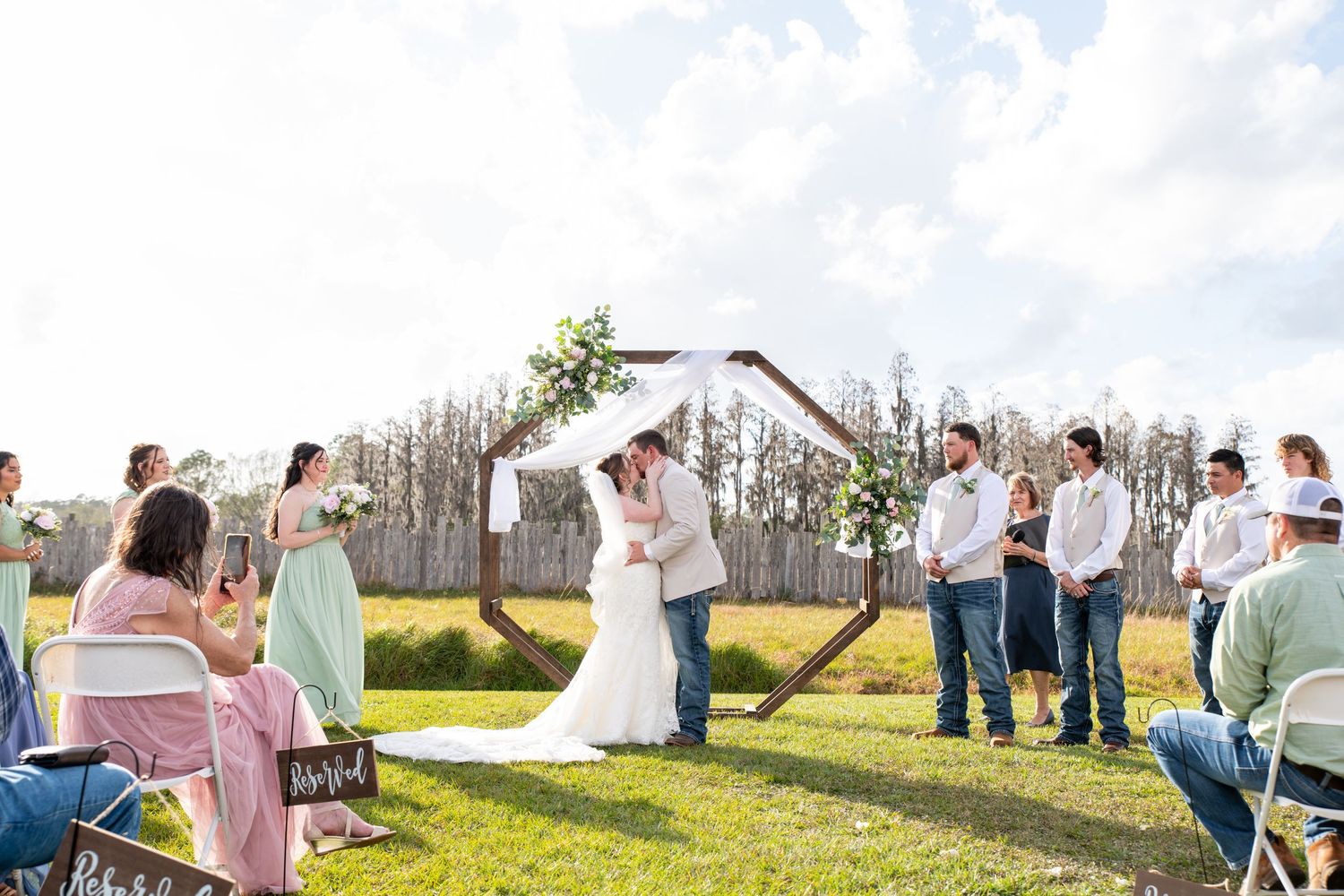 Outdoor wedding ceremony with couple kissing under wooden arch decorated with greenery, surrounded by wedding party and guests.