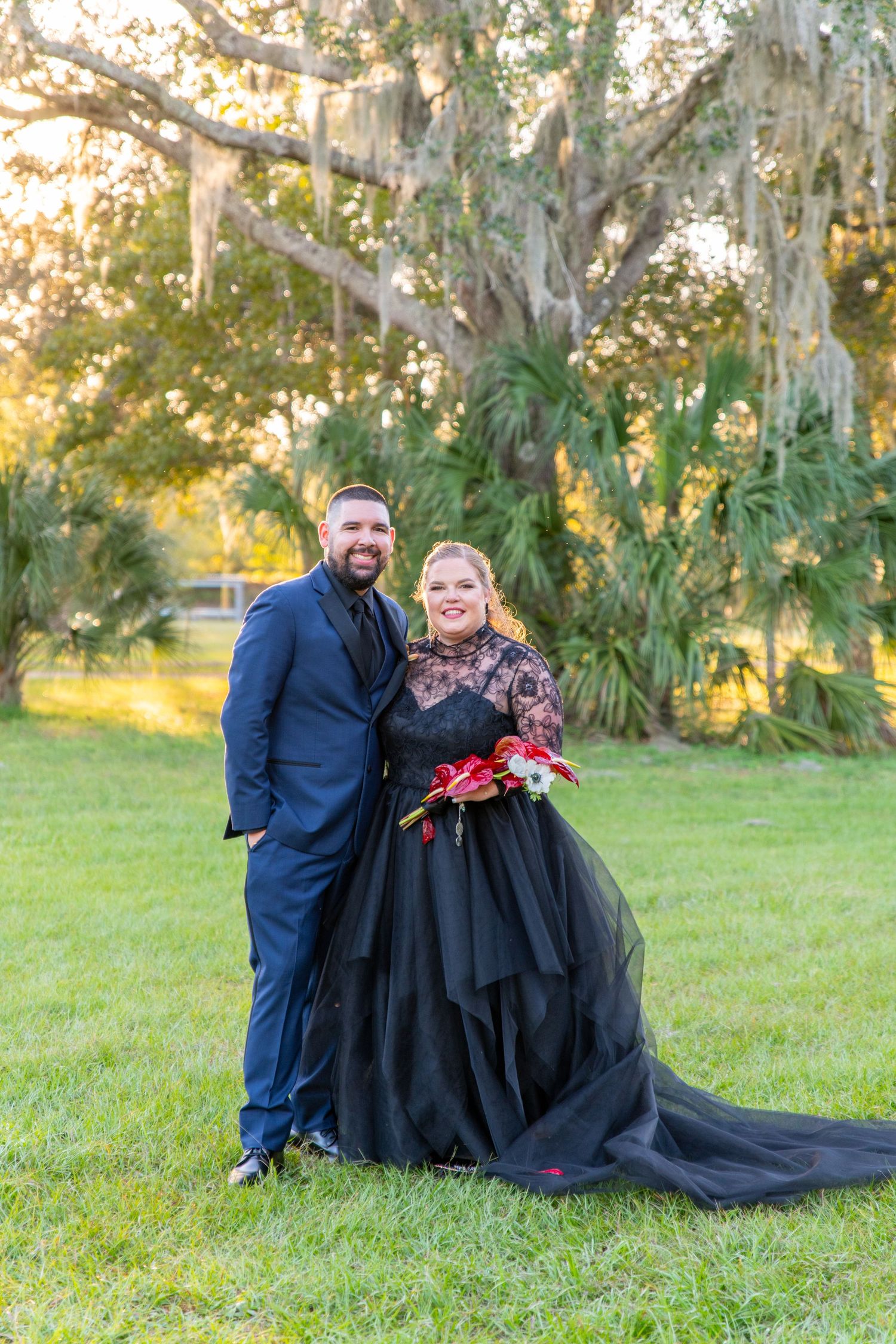 Couple poses outdoors in wedding attire, bride in black gown with red bouquet, groom in navy suit, under Spanish moss trees.