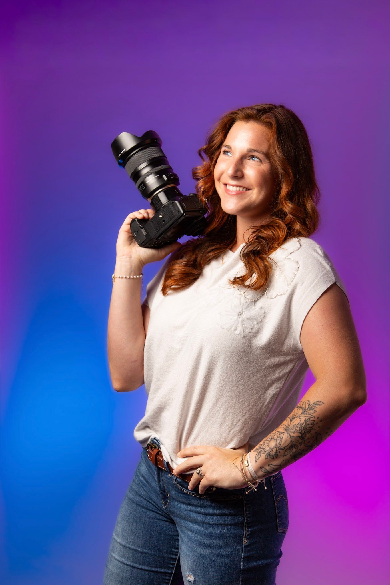 Smiling redhead woman holding a DSLR camera on her shoulder, wearing a white t-shirt and jeans against a purple-blue backdrop.