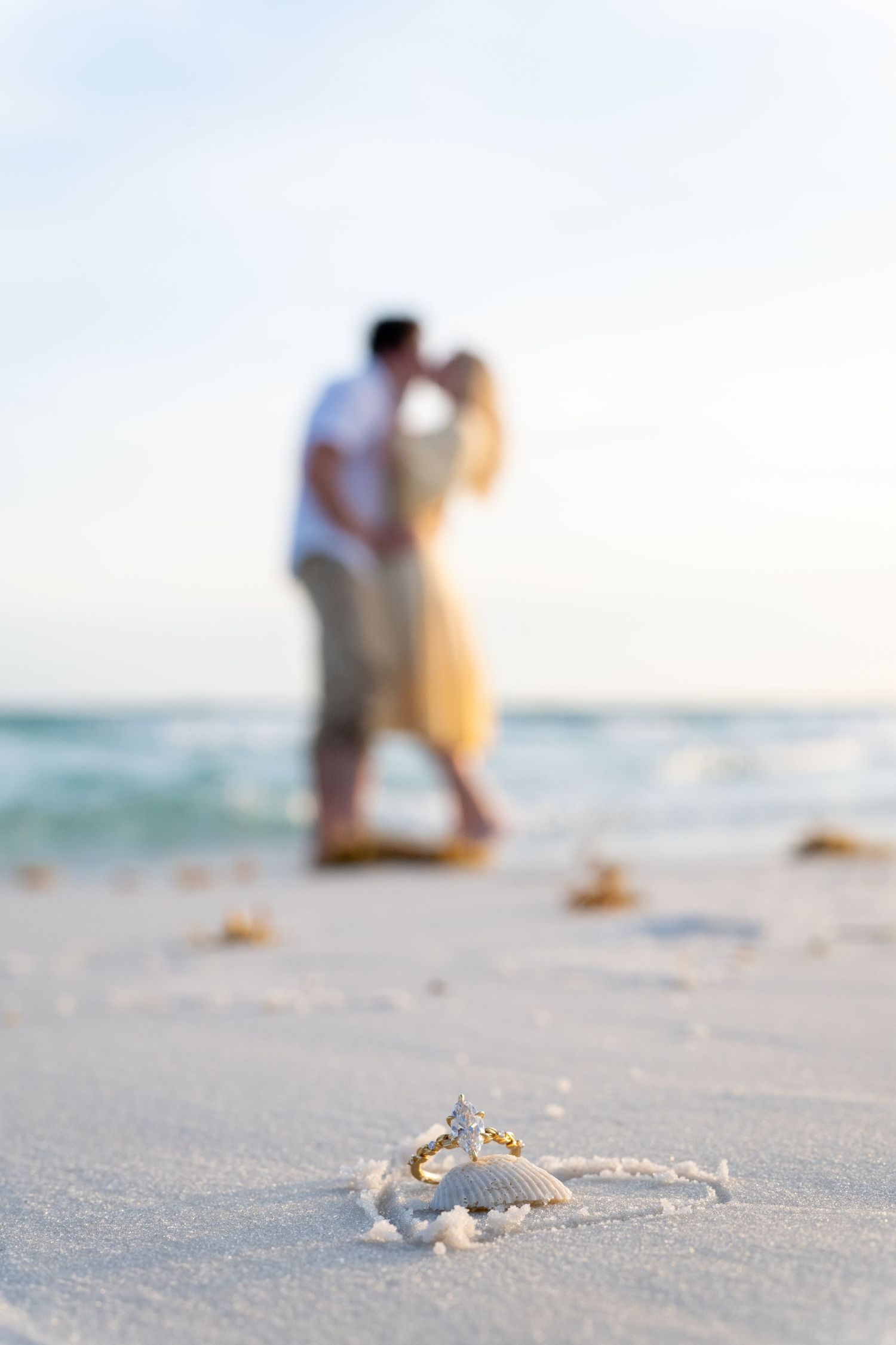 Couple kissing on a beach at sunset with wedding rings on a seashell in the foreground on white sand.