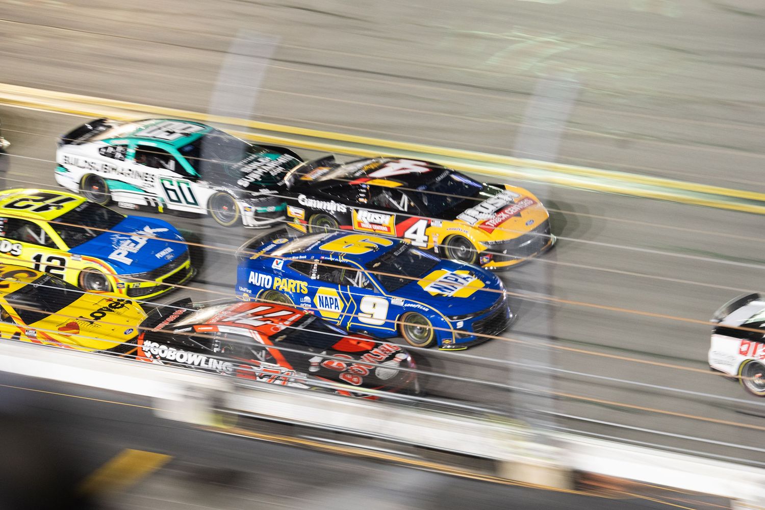Multiple NASCAR race cars racing closely together at high speed during a nighttime event.