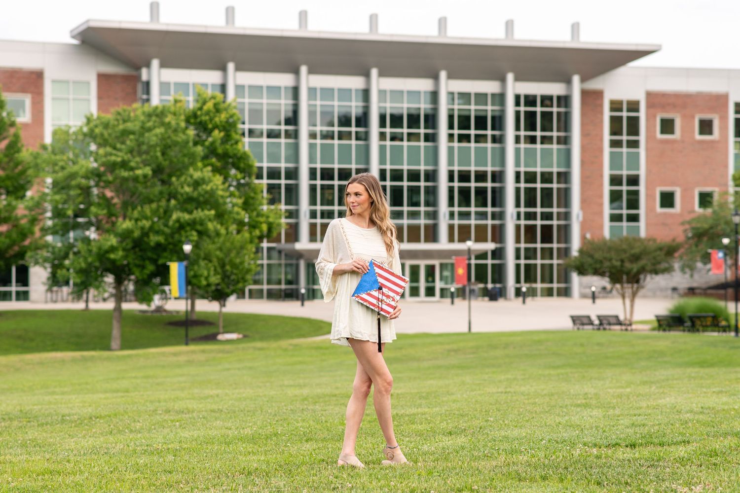 Smiling female graduate in white dress holding American flag on green campus lawn.